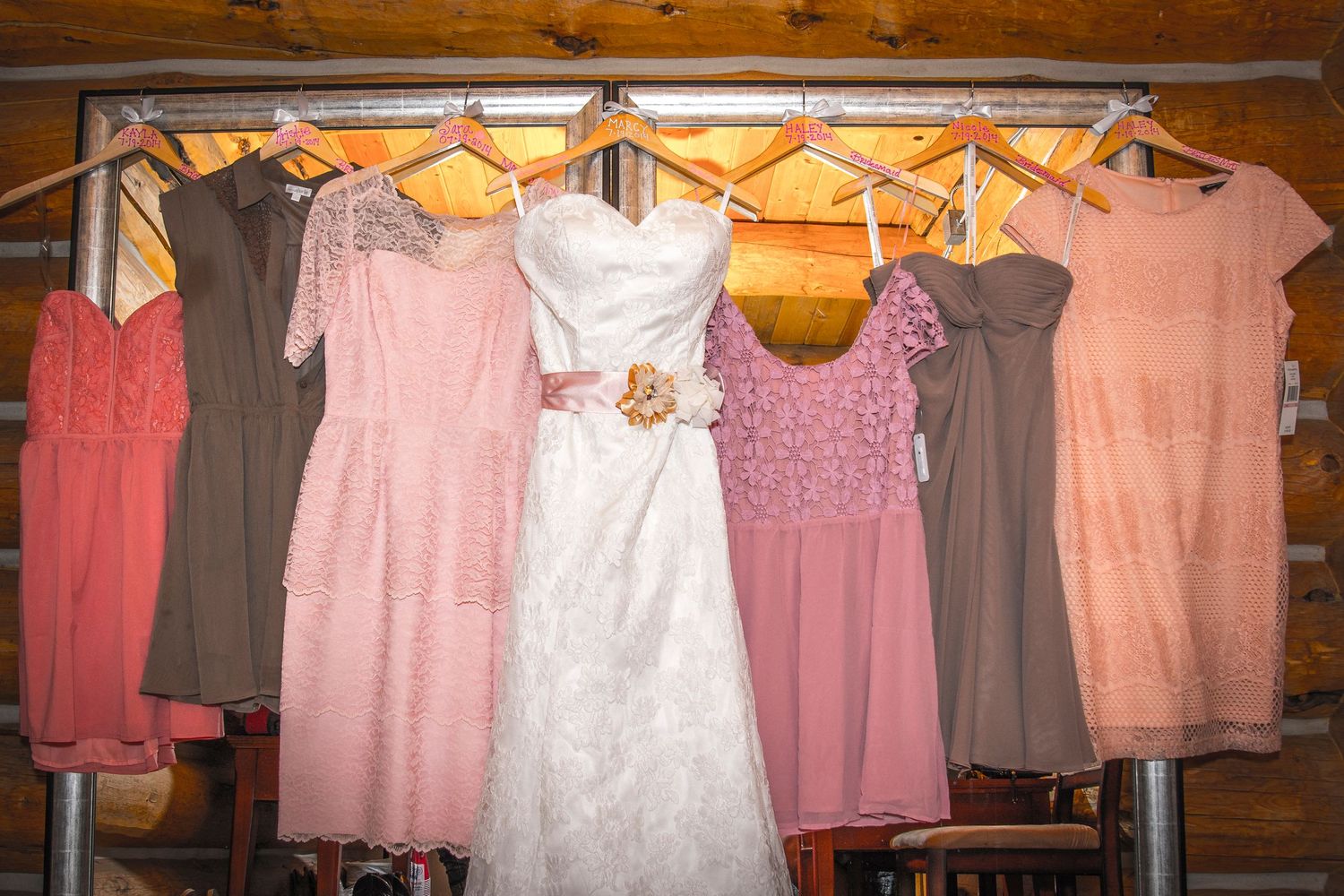 Wedding dress and pink bridesmaid dresses hanging on wooden wall.