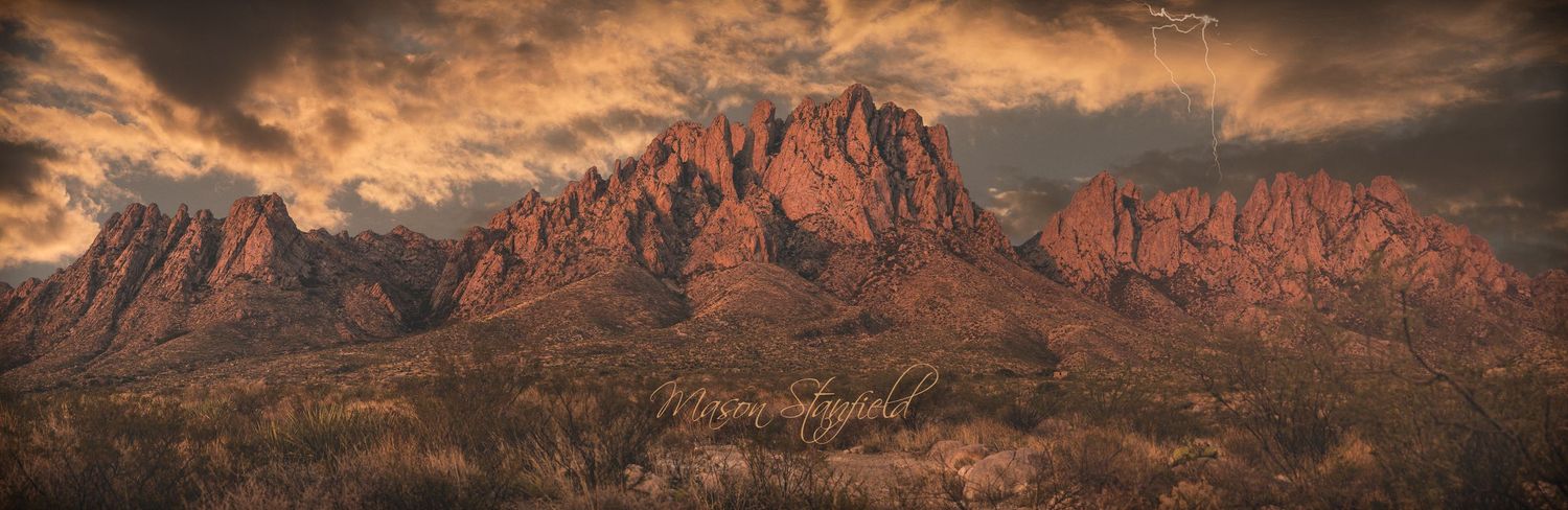 Dramatic sunset sky over red desert mountains with rugged cliffs and desert vegetation in the foreground.