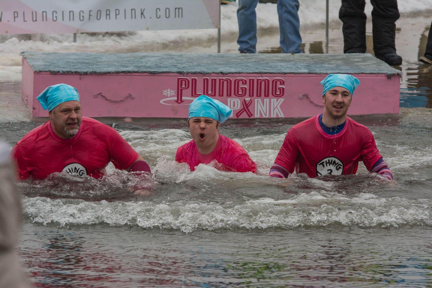 Three participants in pink shirts braving cold water during winter plunge event.