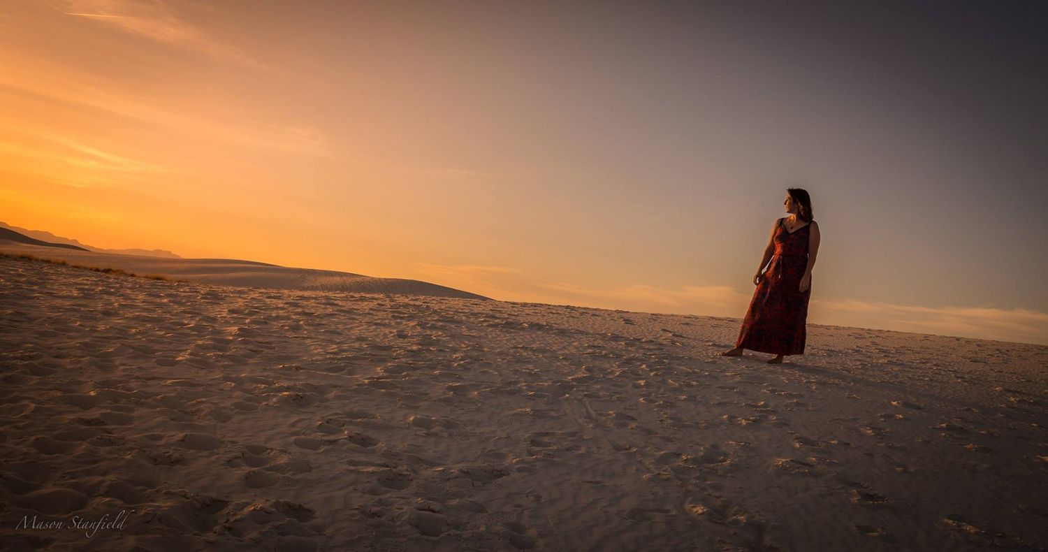 Person in flowing dress stands on sand dune during golden sunset.