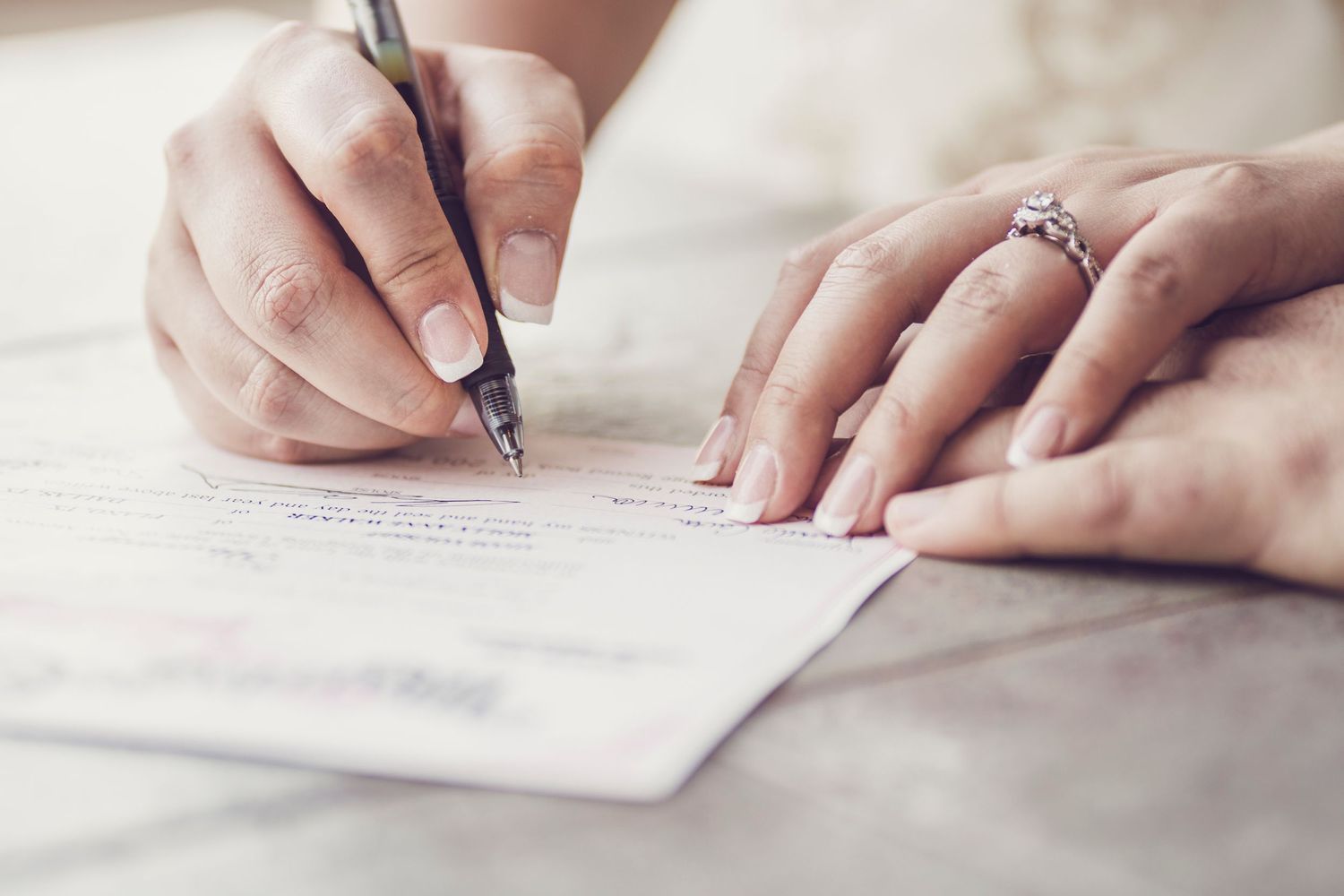 Close-up of hands signing wedding documents with an engagement ring visible.