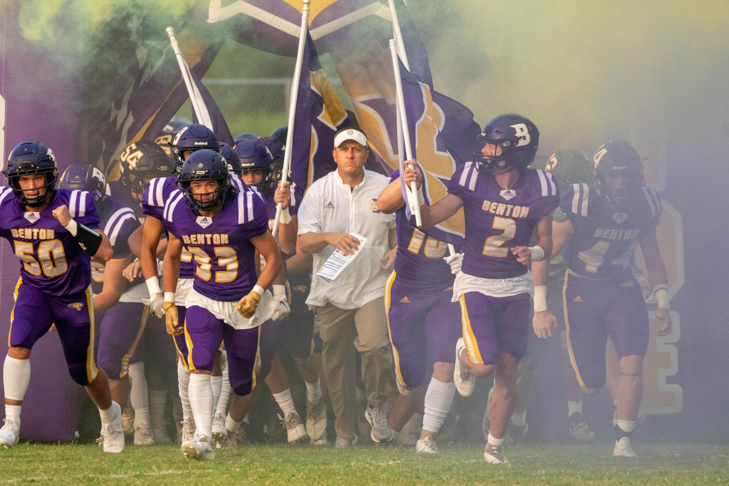 Football team runs onto field through smoke and crossed swords with marching band in background.