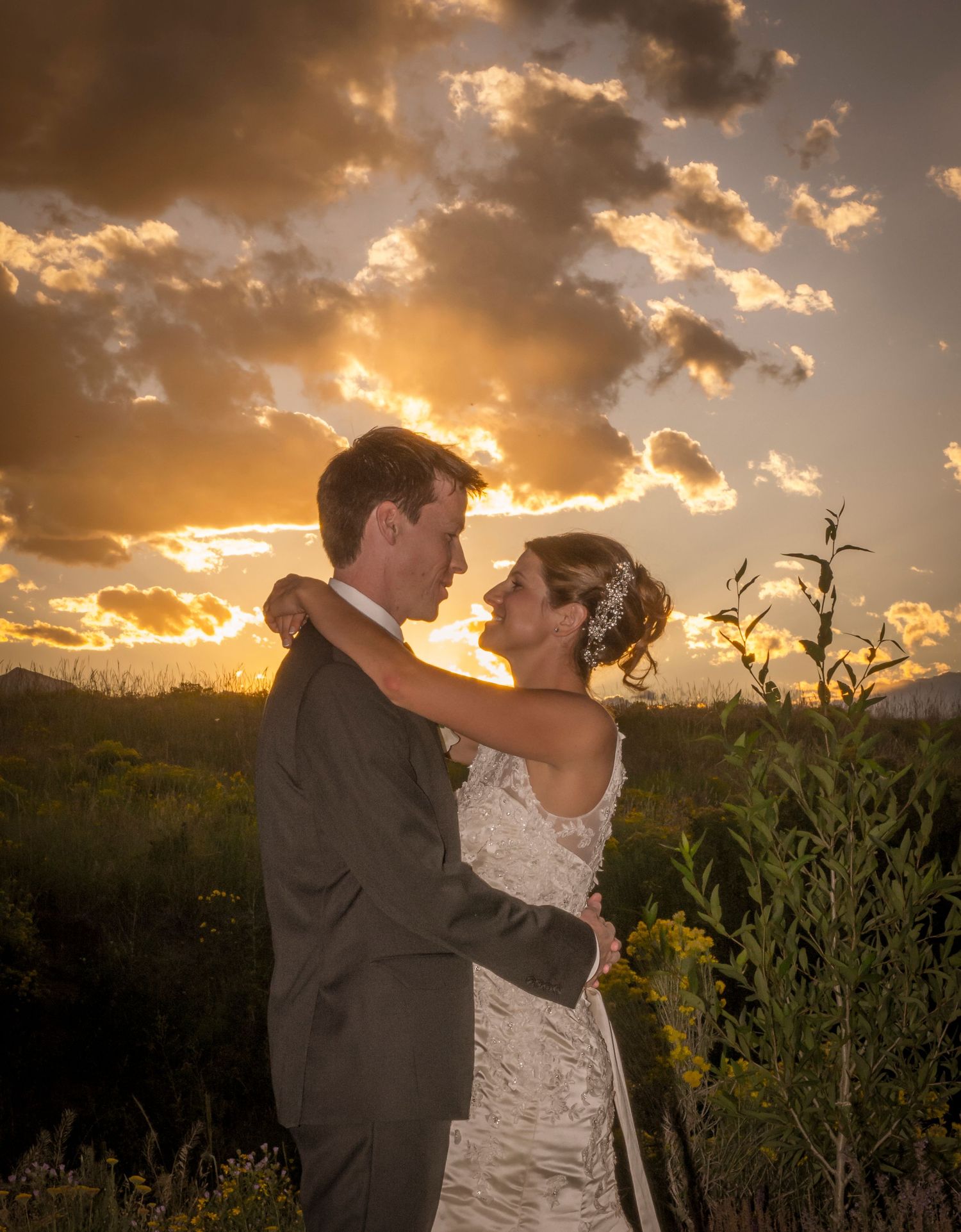 A romantic couple embraces during golden hour with dramatic sunset clouds behind them.