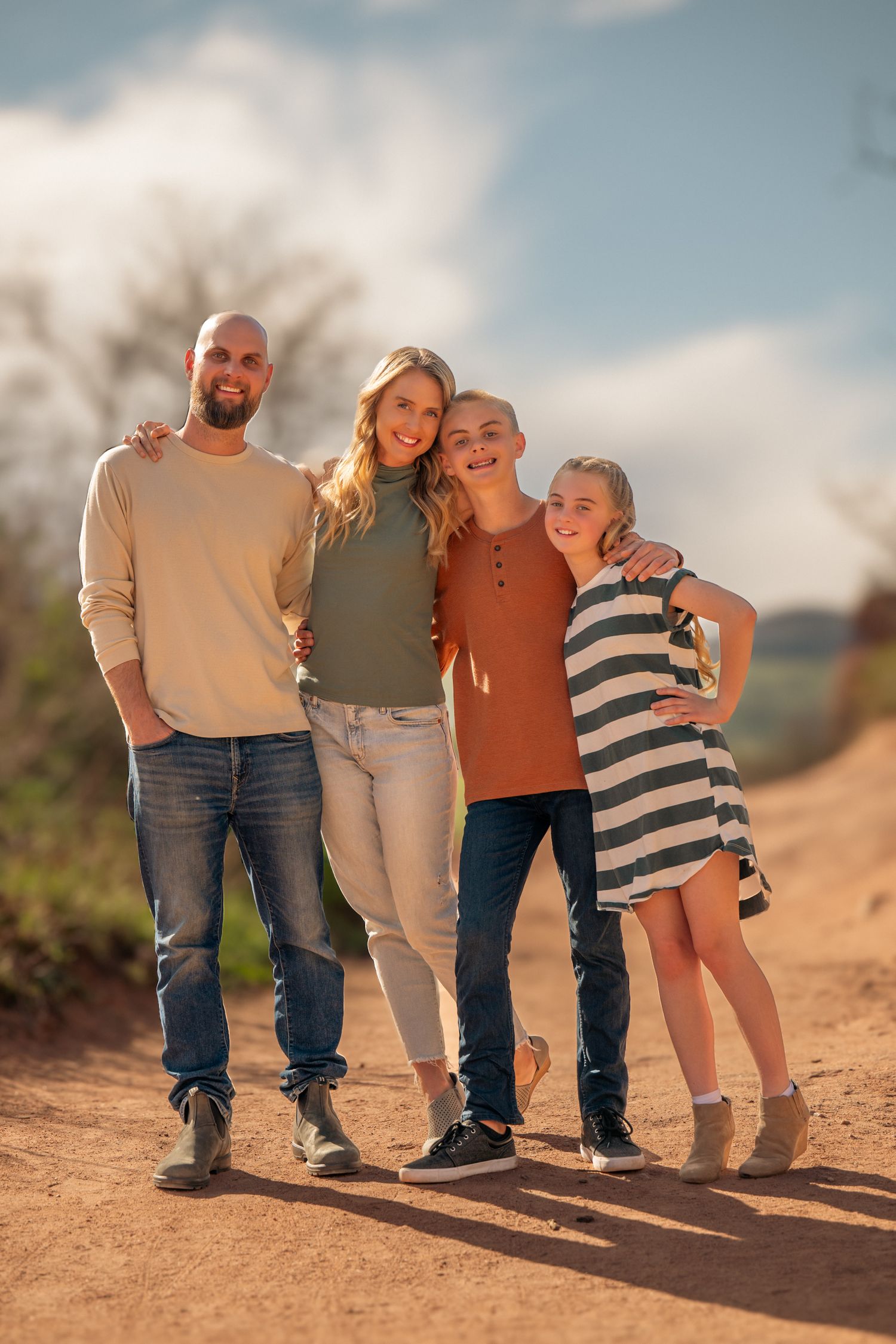 A family of four poses together on a dirt path during golden hour, wearing casual clothing against a cloudy sky.