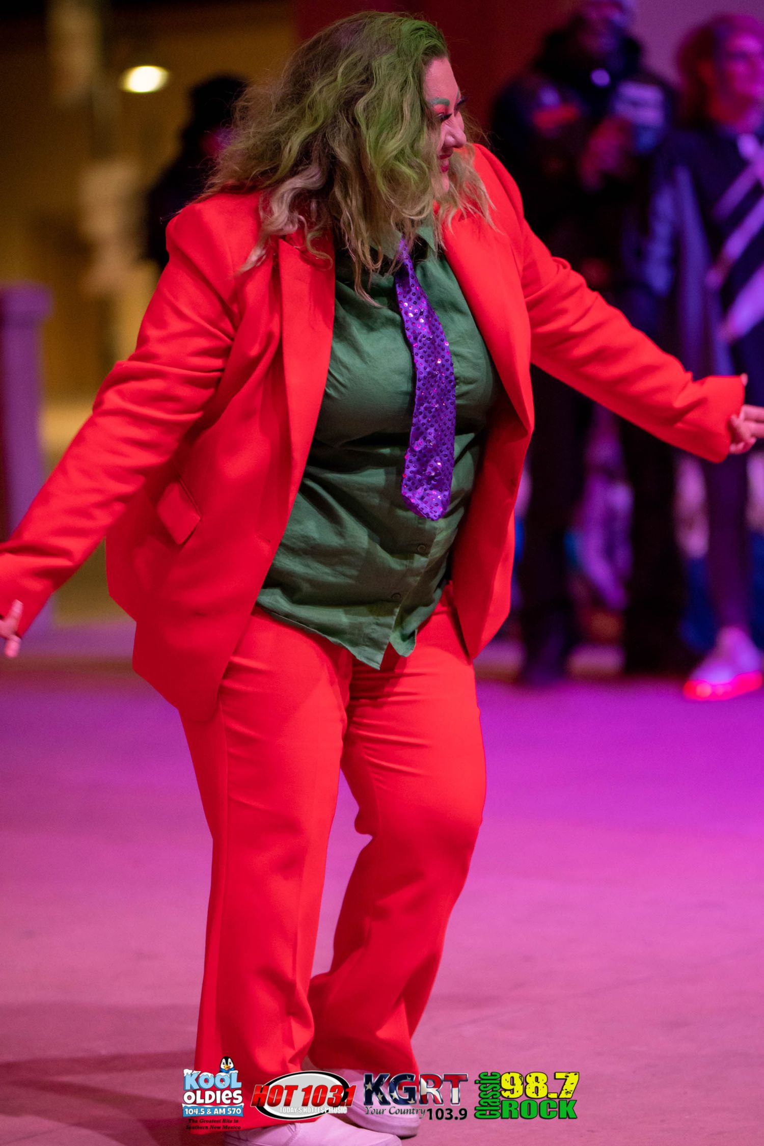 Performer dancing in bright red suit and purple tie under stage lights.