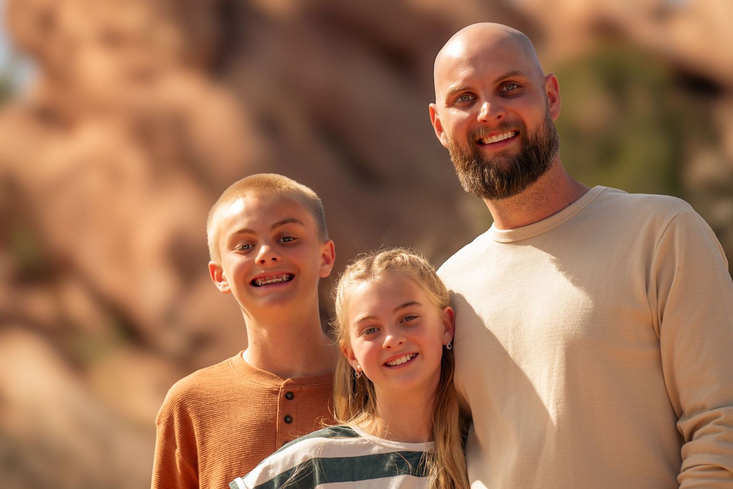 Three family members smile together outdoors against a blurred rocky background in warm sunlight.