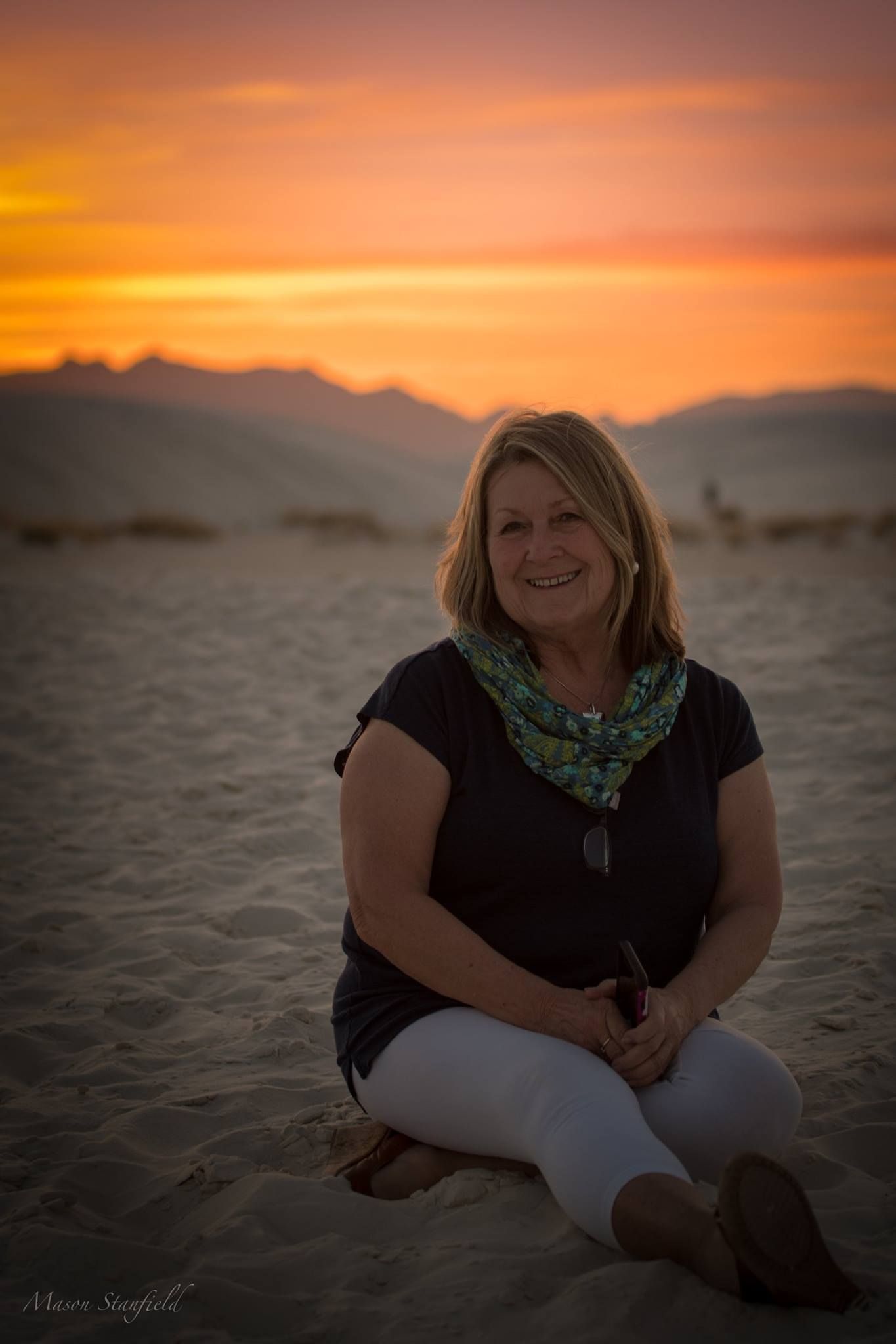 Woman in black shirt and scarf sits on beach sand during vibrant sunset.