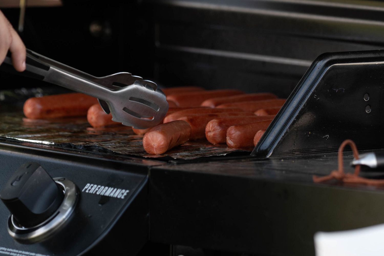 Hot dogs cooking on a black griddle surface with tongs.