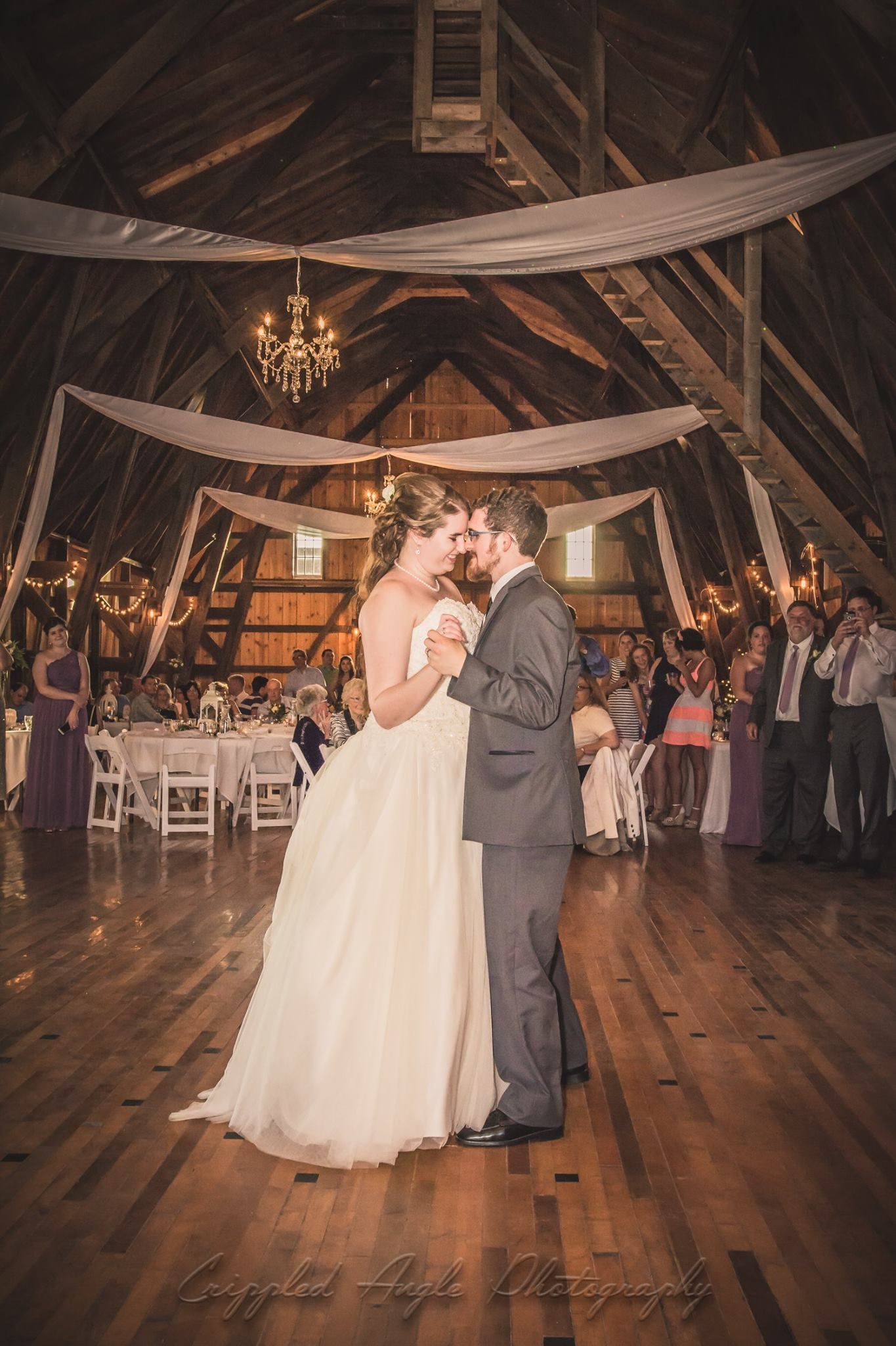 A couple shares their first dance in a rustic barn venue with draped fabric and chandelier decor.