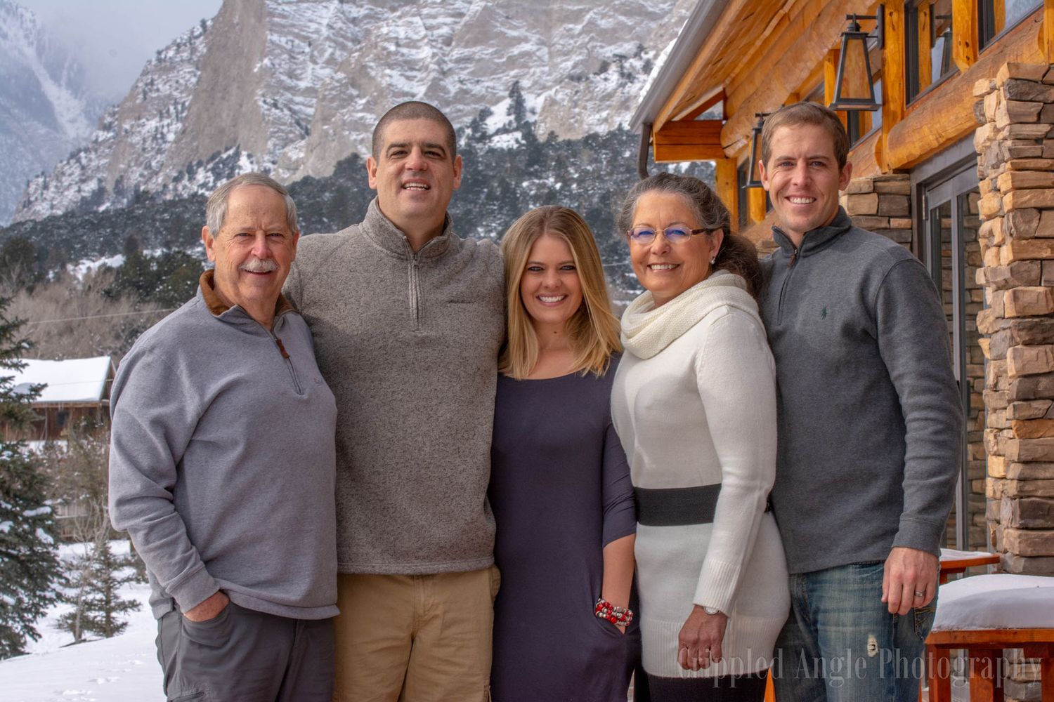 A family group poses together outside a mountain cabin in winter with snowy peaks behind them.