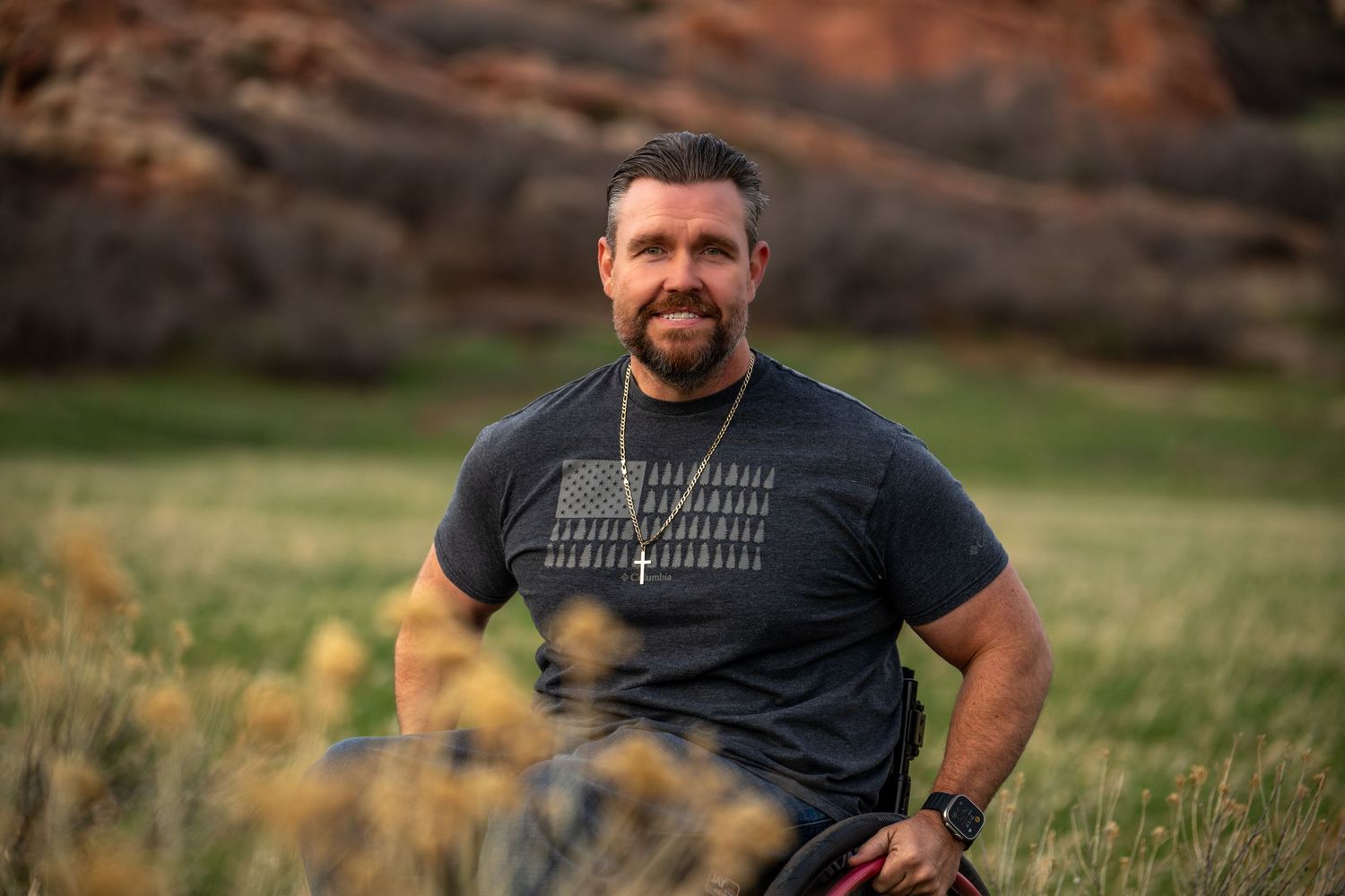 A person poses outdoors wearing a black t-shirt in a grassy field with mountainous terrain in the background.