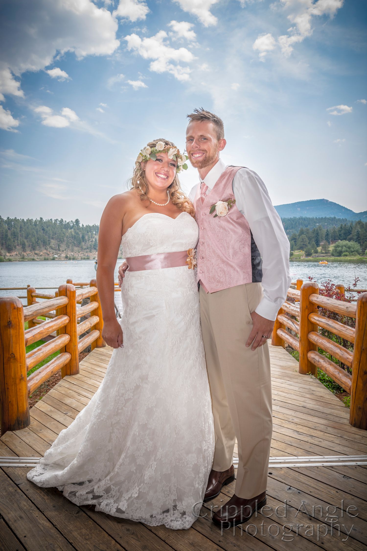 Newlyweds pose on wooden deck overlooking mountain lake on their wedding day.