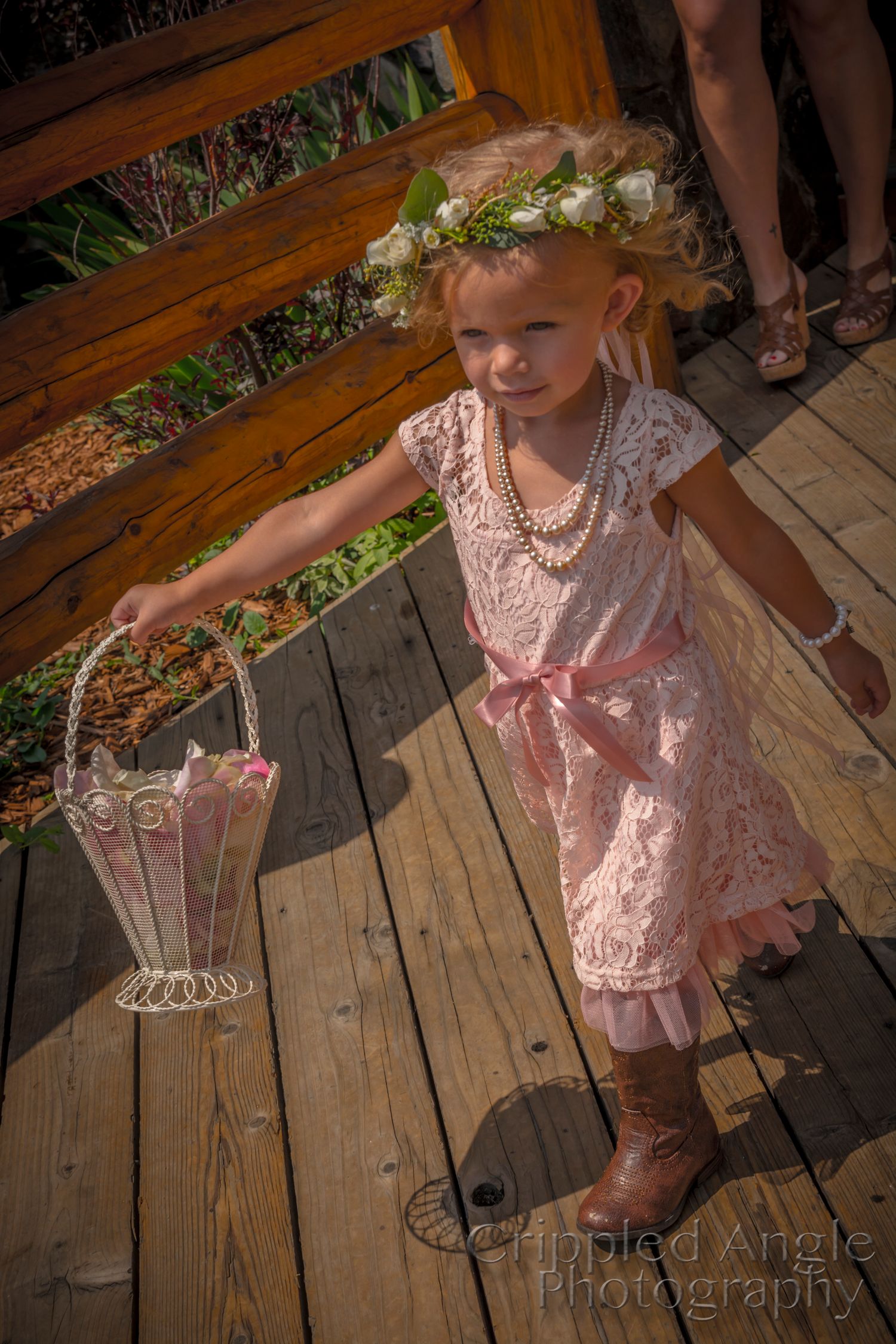 Young flower girl in pink lace dress and floral crown holds a decorative basket on wooden deck.
