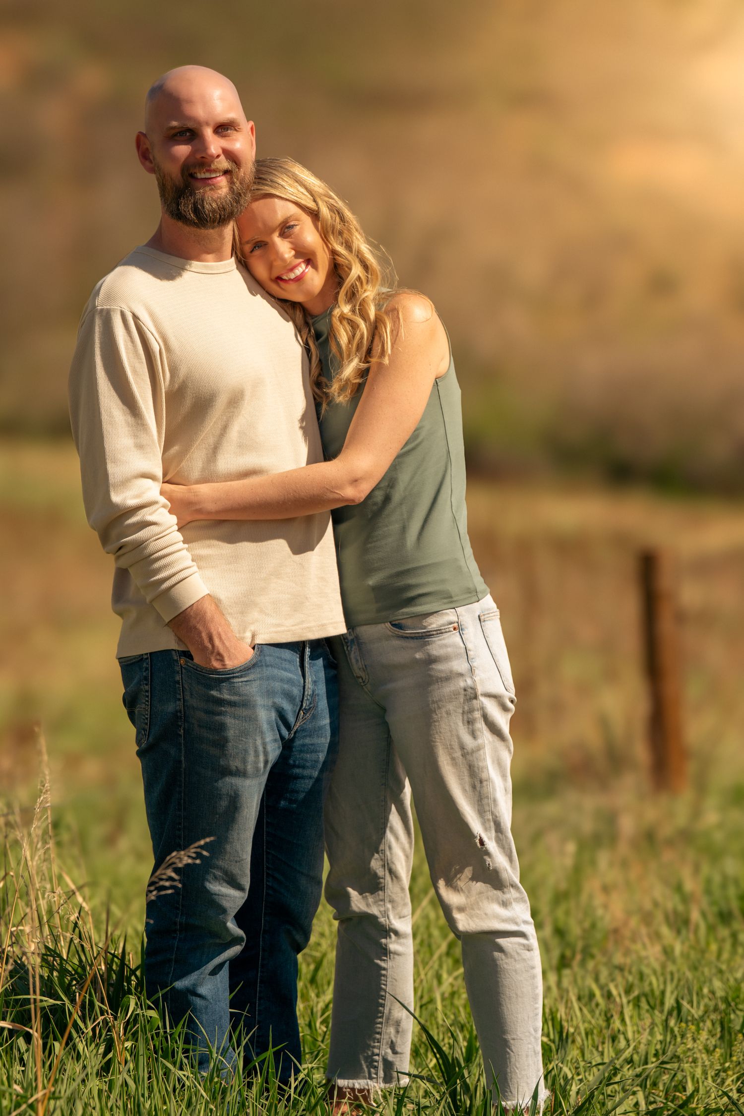 A couple embraces in a sunlit meadow with golden light and mountains in the background.