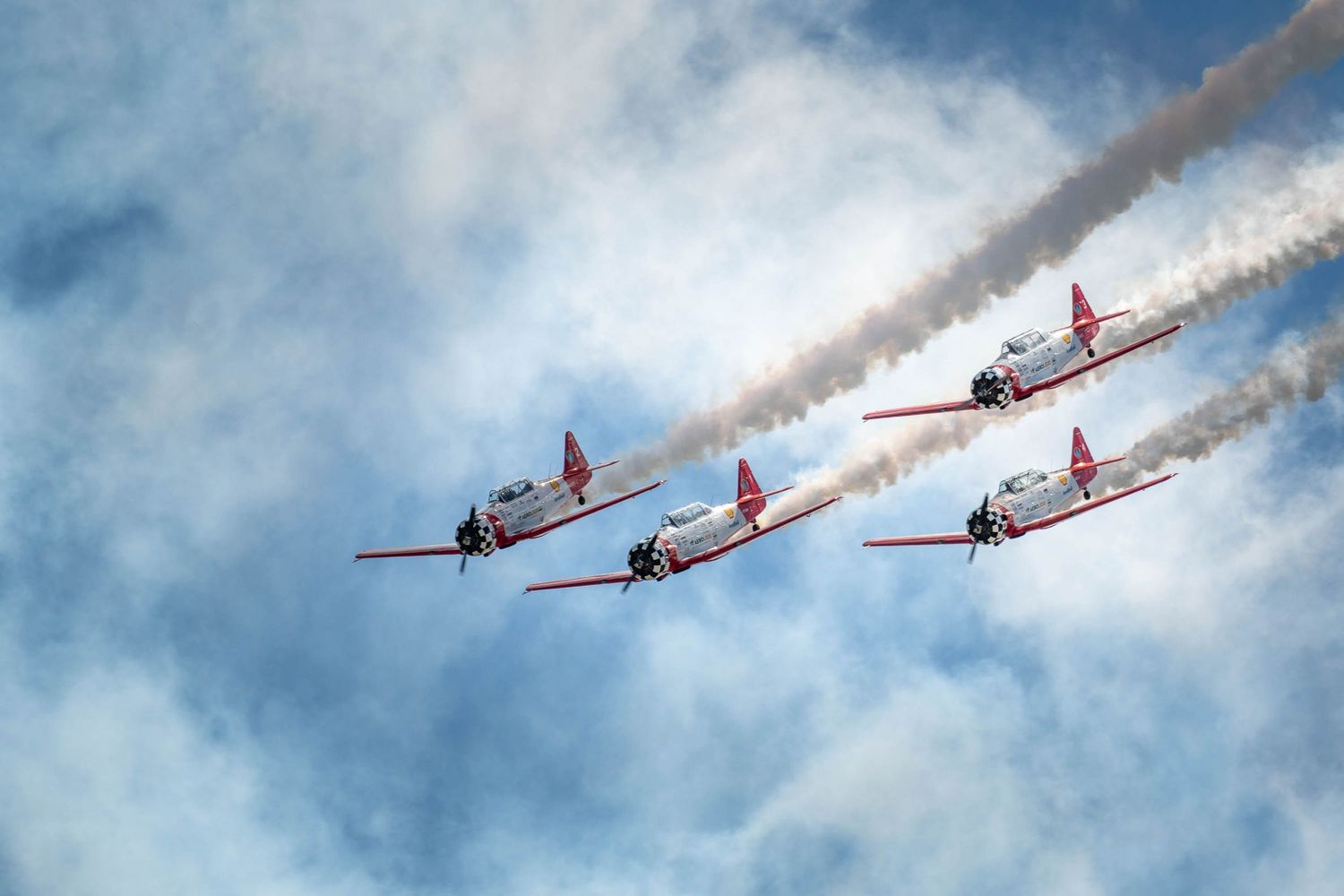 Formation of red vintage aircraft performing an airshow with white smoke trails against blue sky.