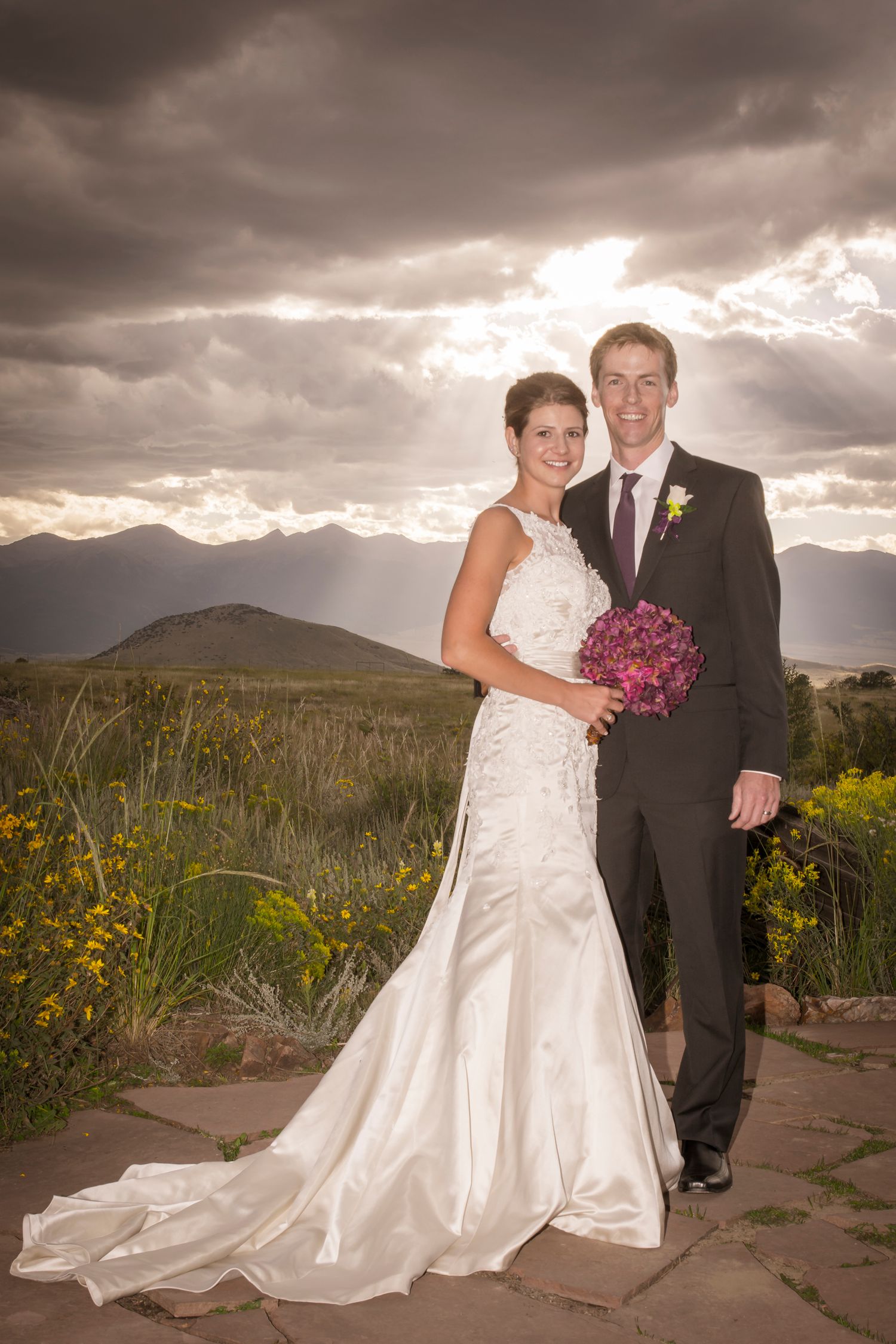 Newlyweds pose for portrait with dramatic mountain landscape and stormy sky.