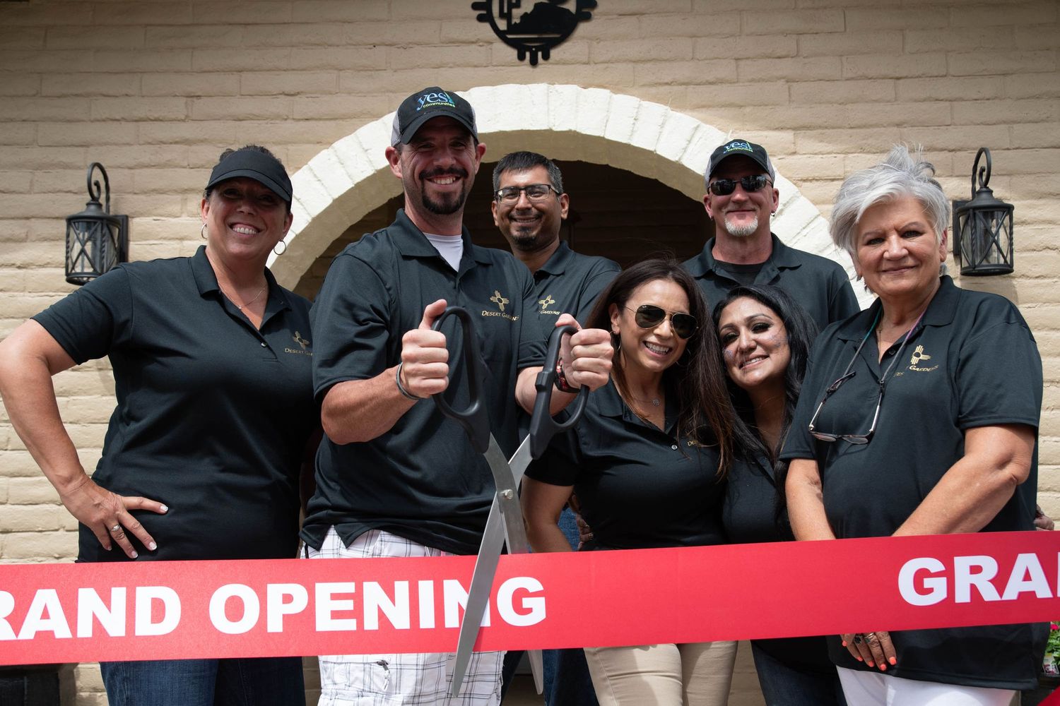 Group celebrating at grand opening with red ribbon and thumbs up gestures.