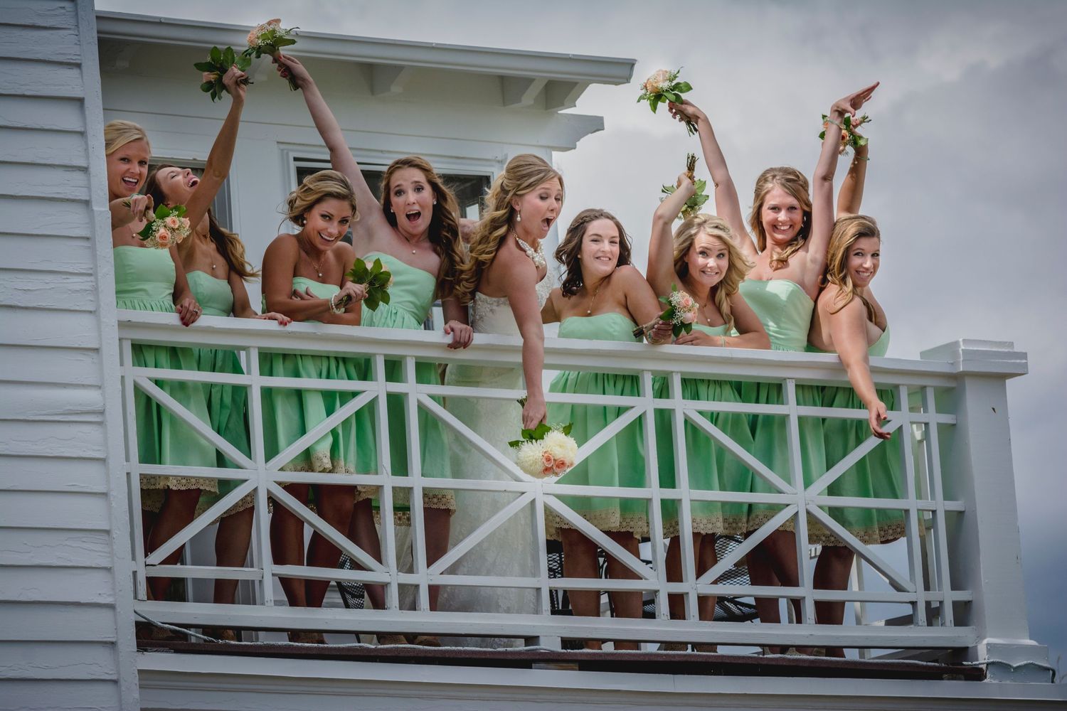 Bridesmaids in mint green dresses gleefully pose together on a white porch railing during a wedding celebration.