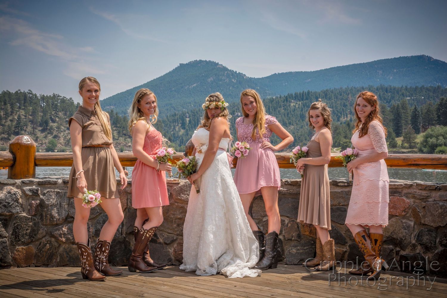 Bridal party in pink and beige dresses pose with cowboy boots against mountain backdrop.