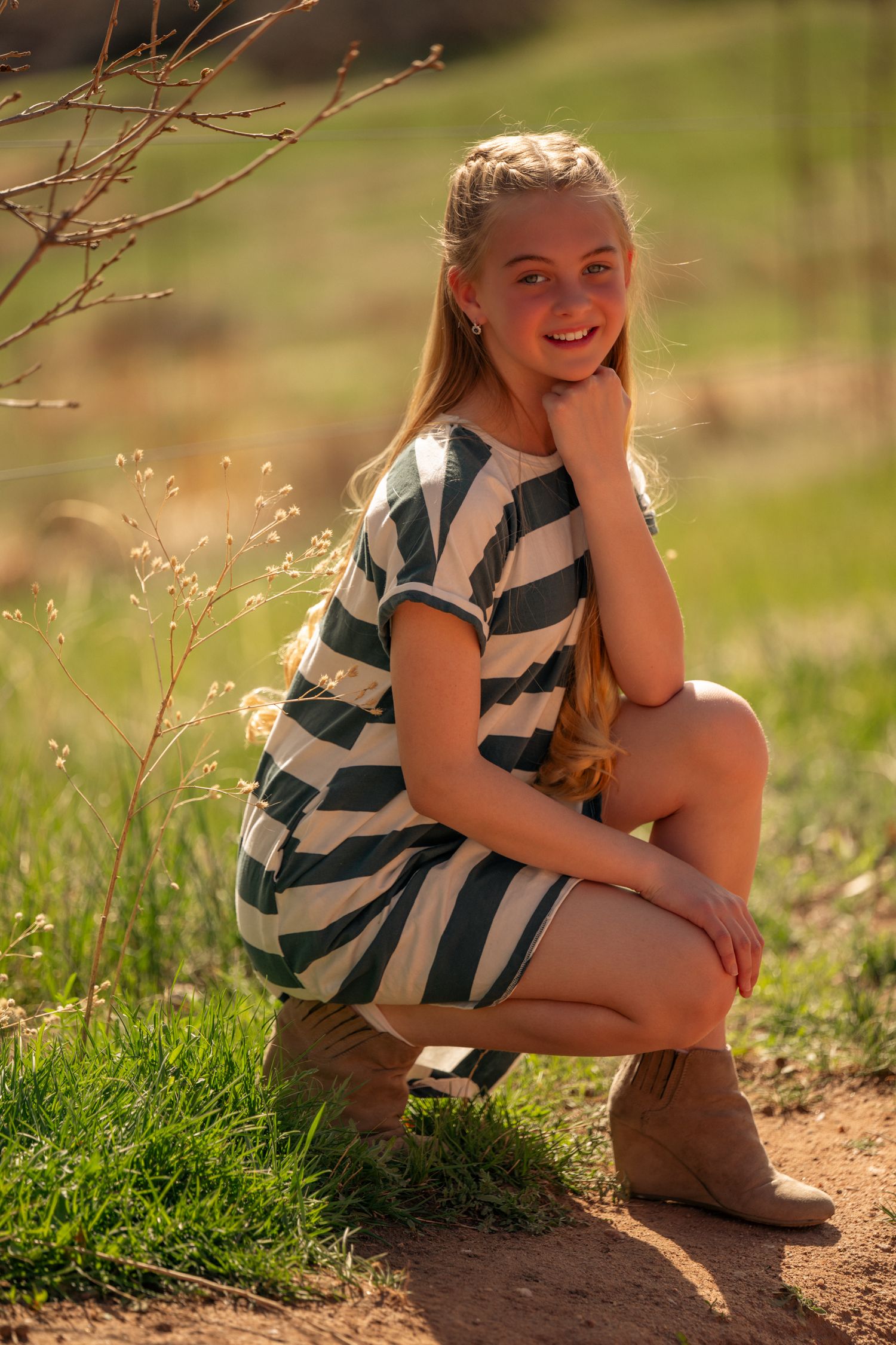 A young person in a striped dress crouches on a dirt path during golden hour light while wearing brown boots.