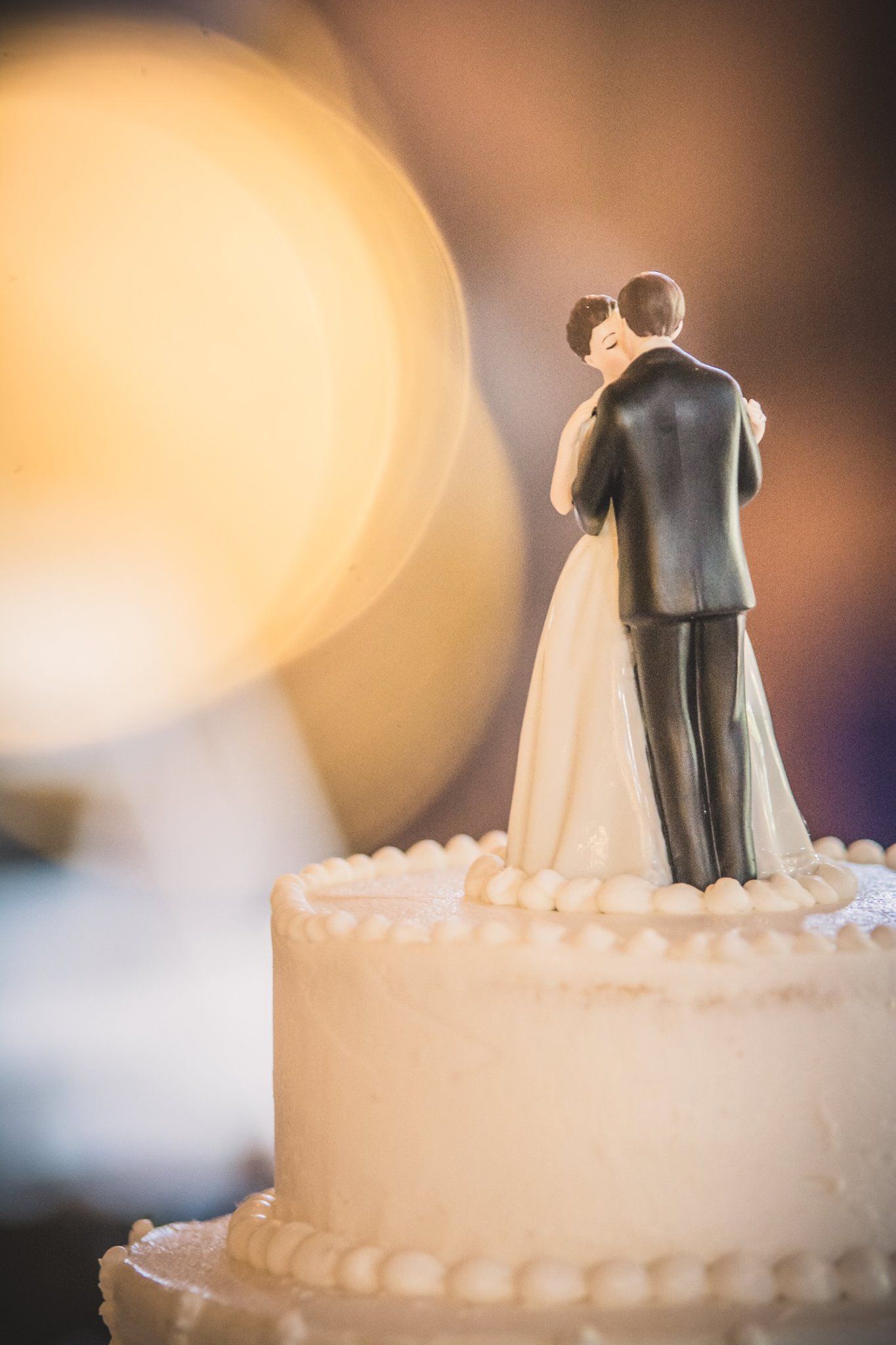 A classic wedding cake topper silhouette stands atop a white frosted cake with bokeh lighting.