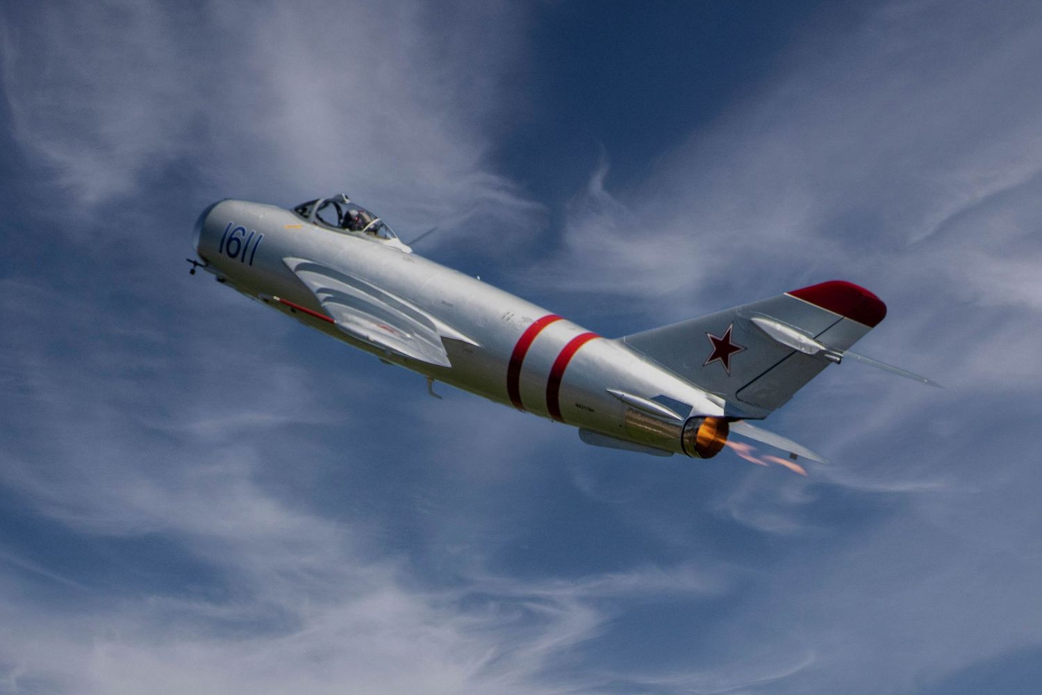 Vintage silver fighter jet with red stripes performs aerial maneuver against cloudy sky.