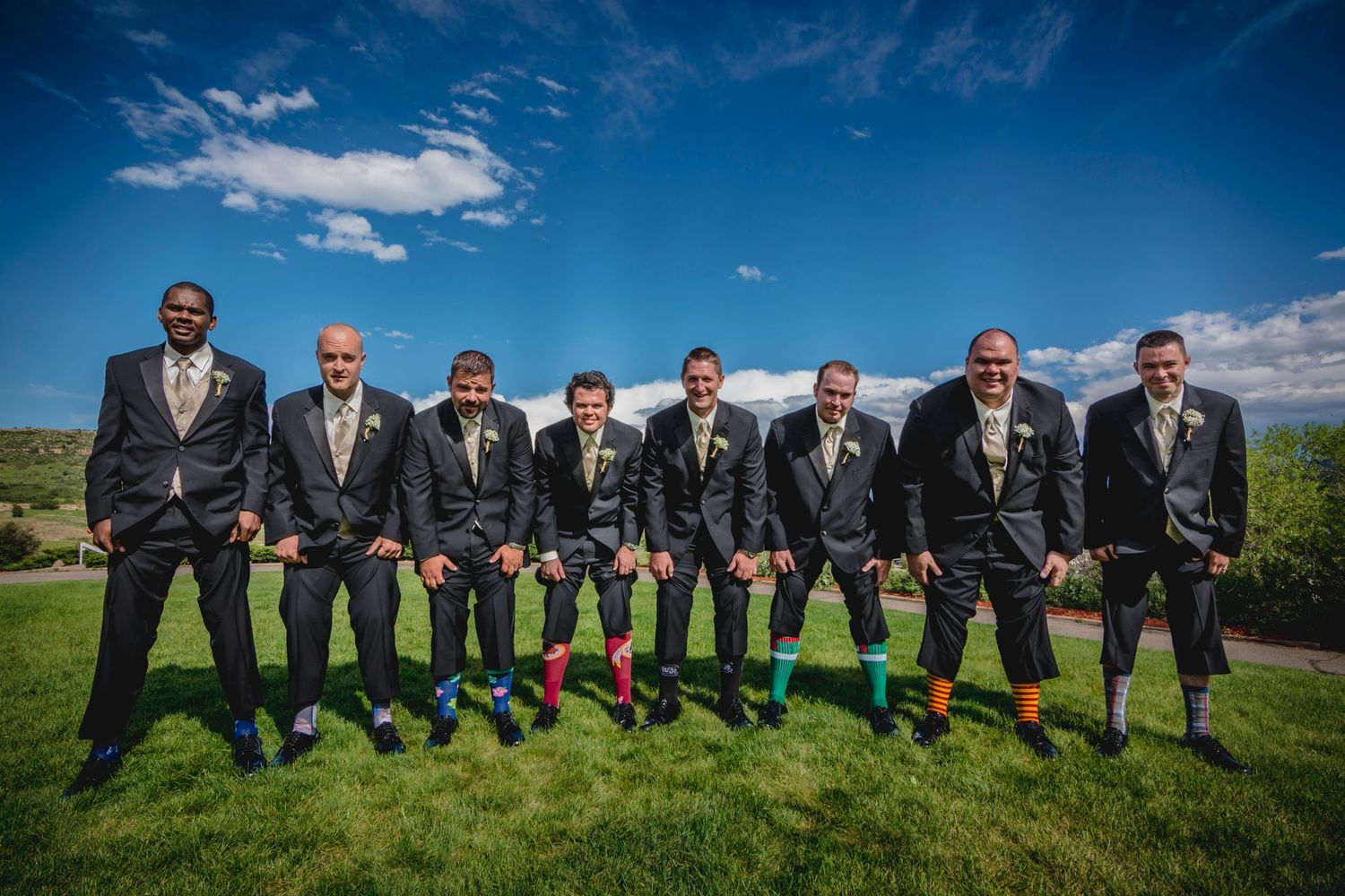 Groomsmen wearing black suits with bright green socks pose together outdoors under blue skies.