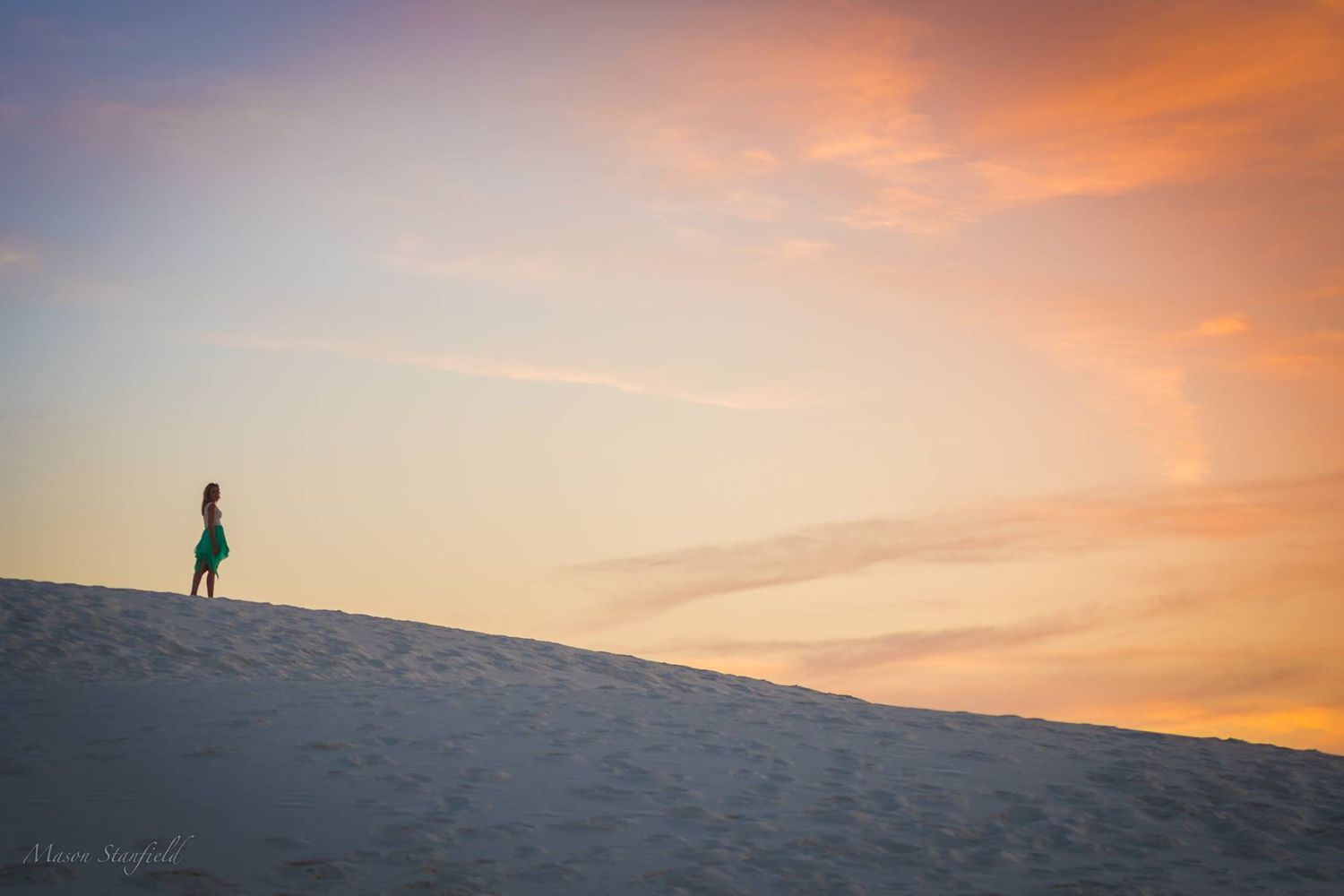 Silhouette of person standing on sand dune during orange sunset.