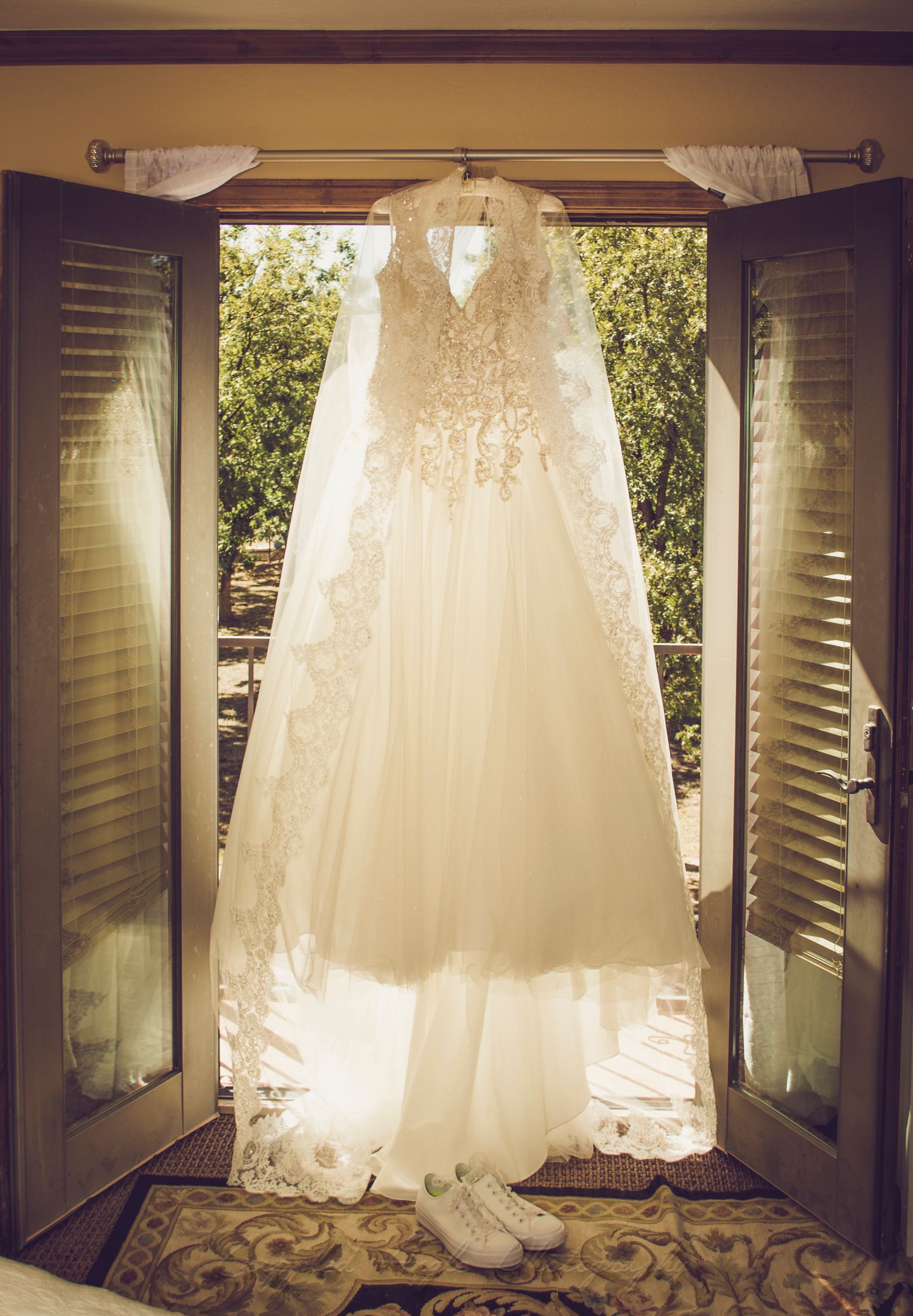 A beautiful white lace wedding dress hanging in a window with natural sunlight streaming through.