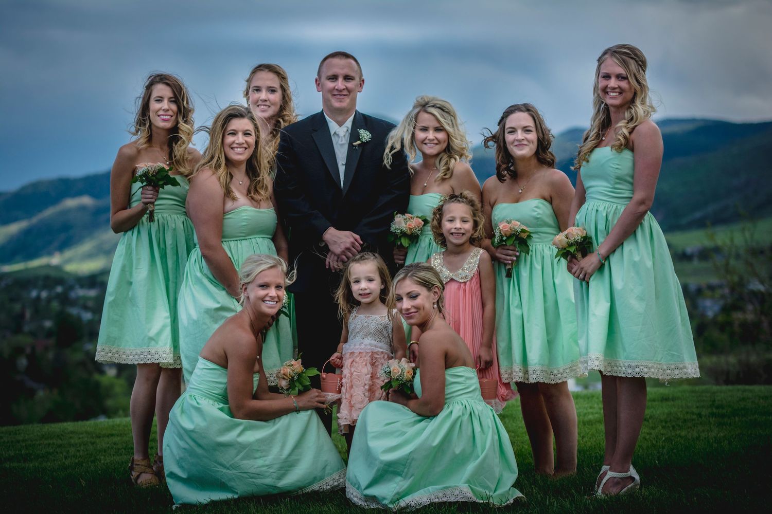 Wedding party in mint green dresses poses on a grassy hillside with mountains in the background.