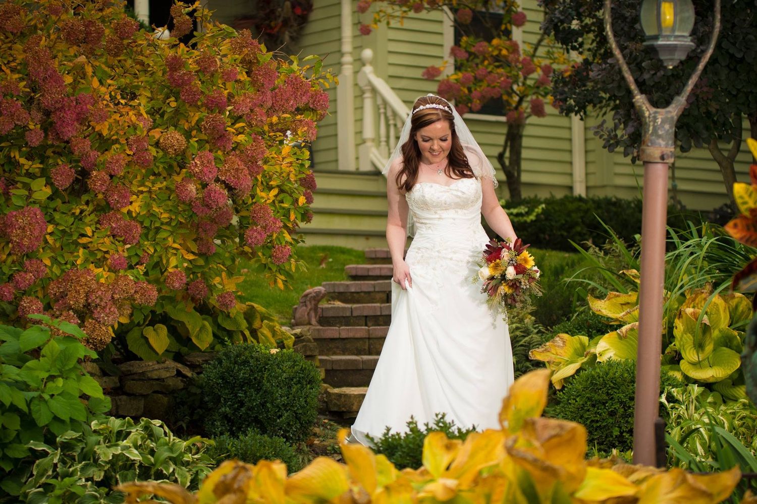 A bride in white dress descends garden steps surrounded by vibrant fall foliage.