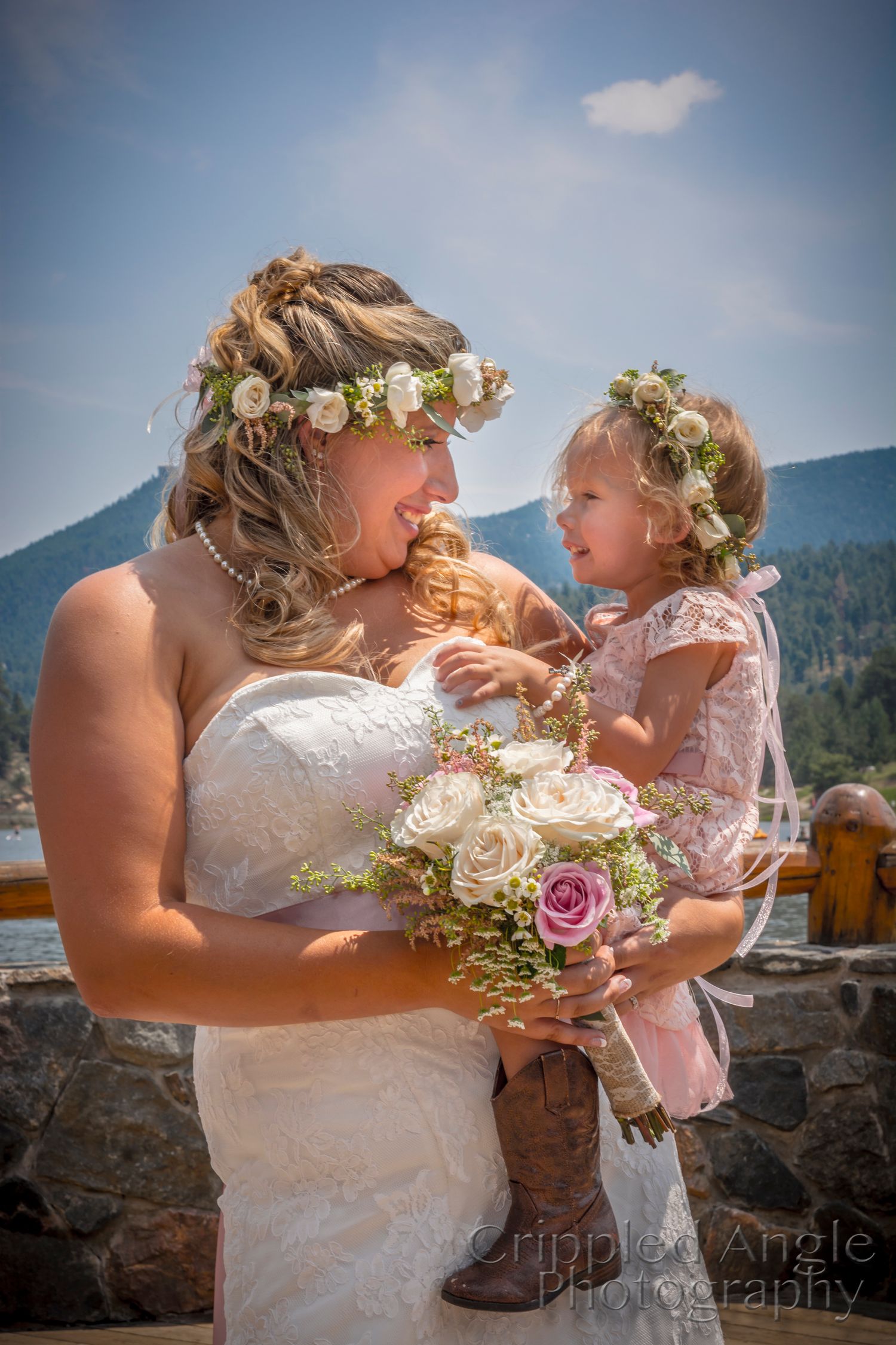 Bride in white dress and floral crown shares tender moment with young flower girl.