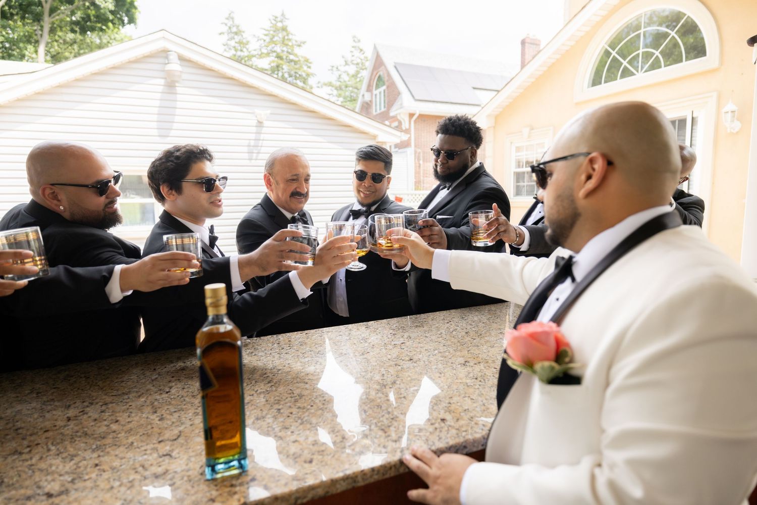 Wedding party members in black and white formal wear toast with beer bottles at an outdoor venue.