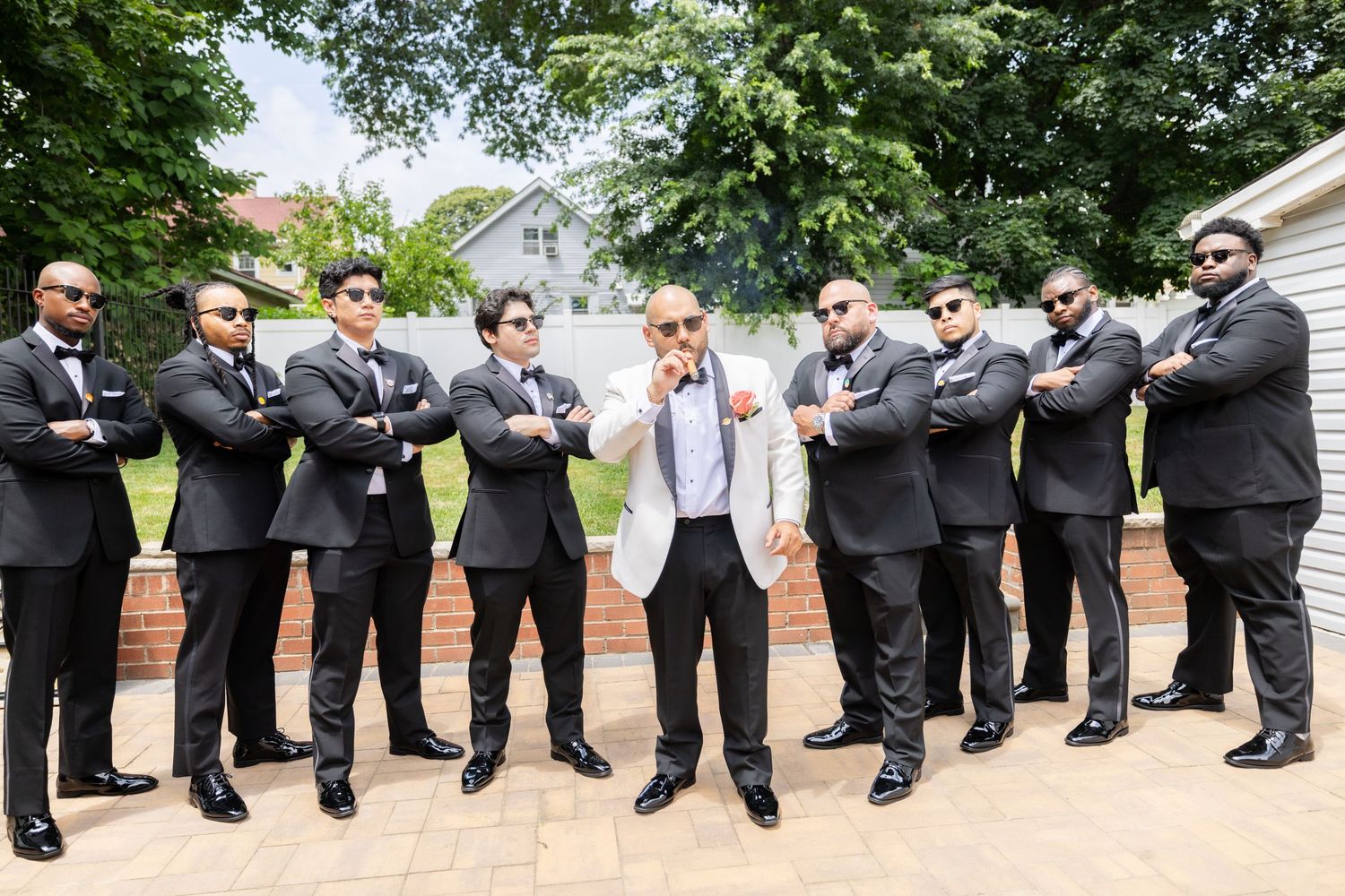Groomsmen in black suits strike a fun pose while standing in a curved line outdoors during a wedding.