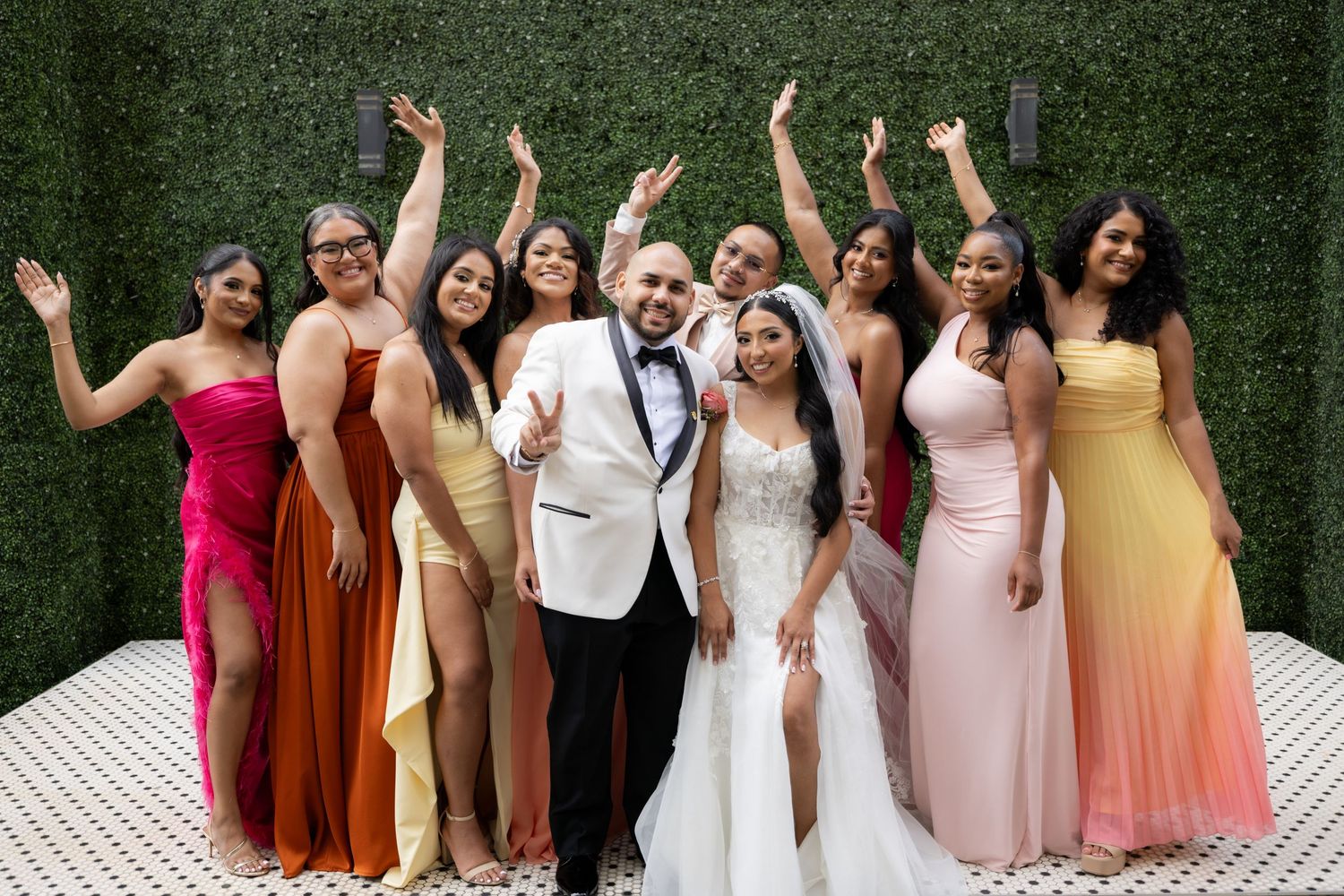 Wedding party poses together outdoors against a green hedge wall wearing coordinated dresses in warm autumn tones.
