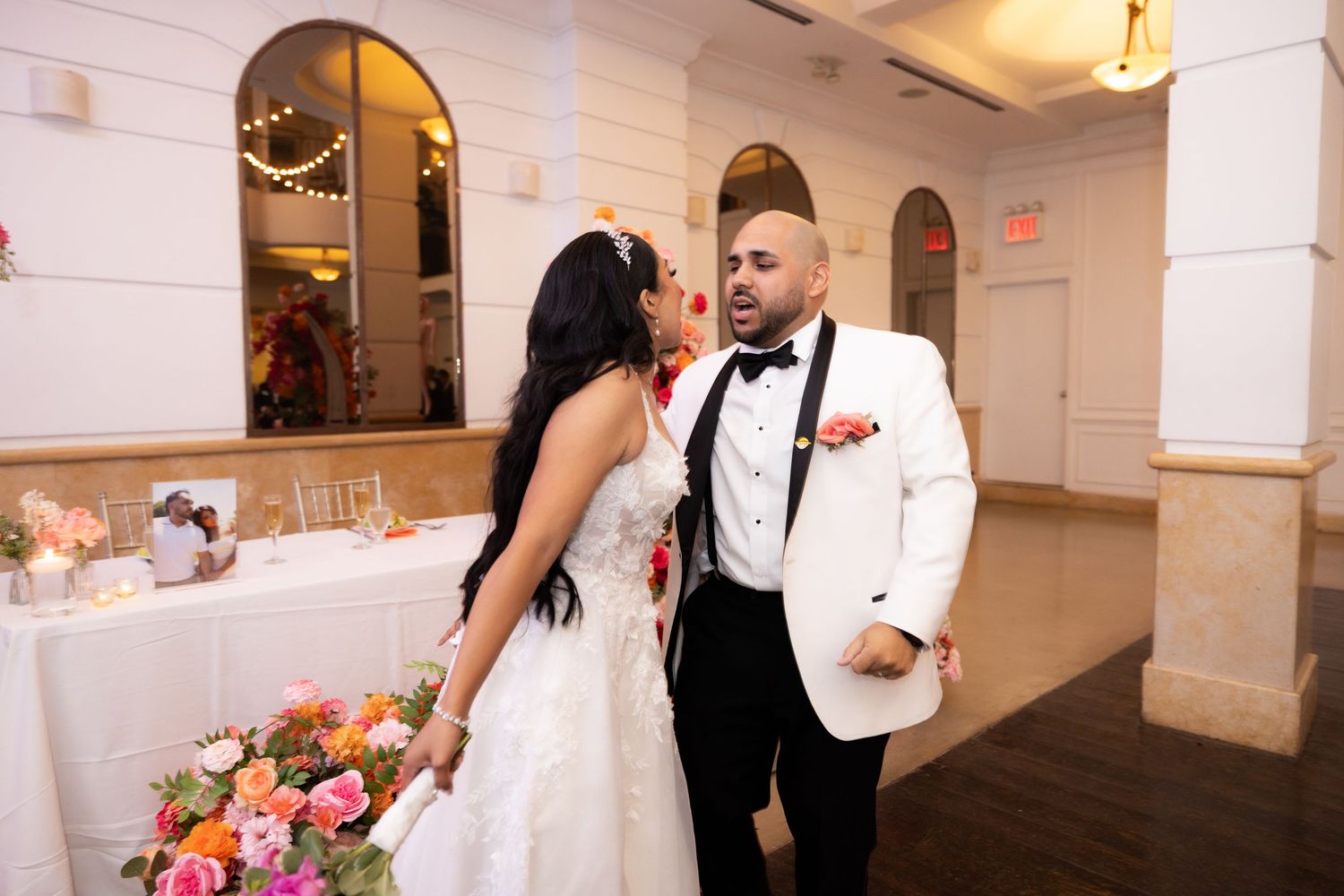 Wedding party dancing and celebrating in a white-walled reception venue with decorative lighting.