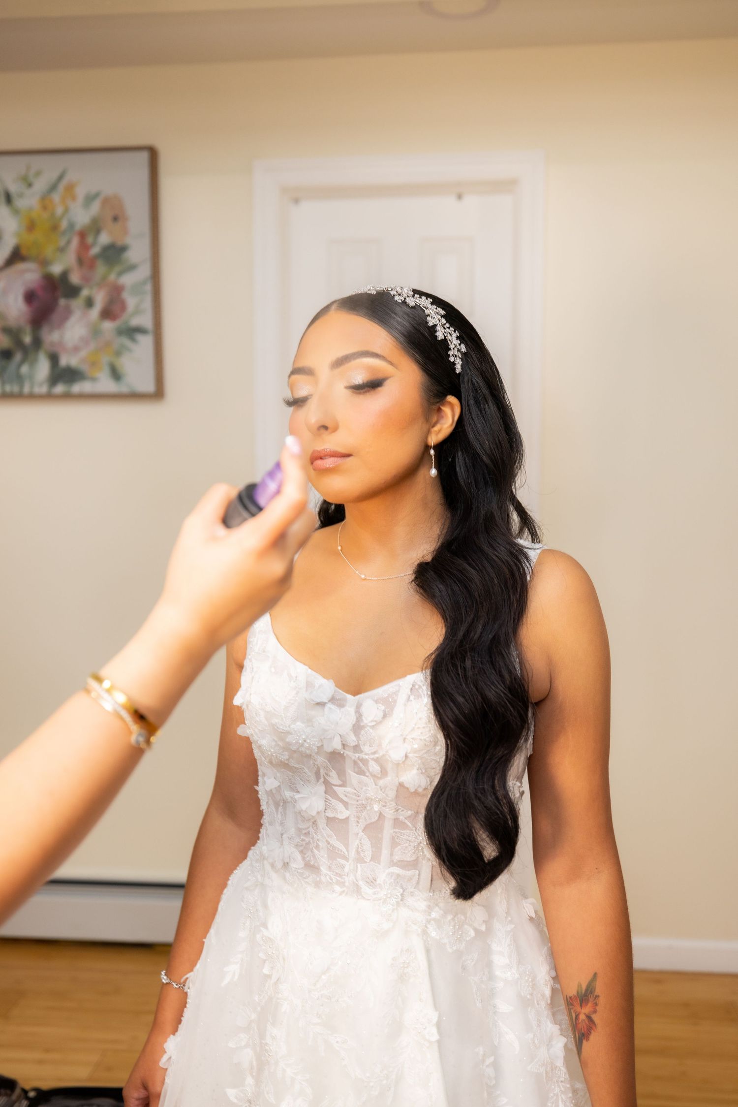 A bride wearing a white lace wedding gown with long dark hair styled in glamorous waves poses during preparation.