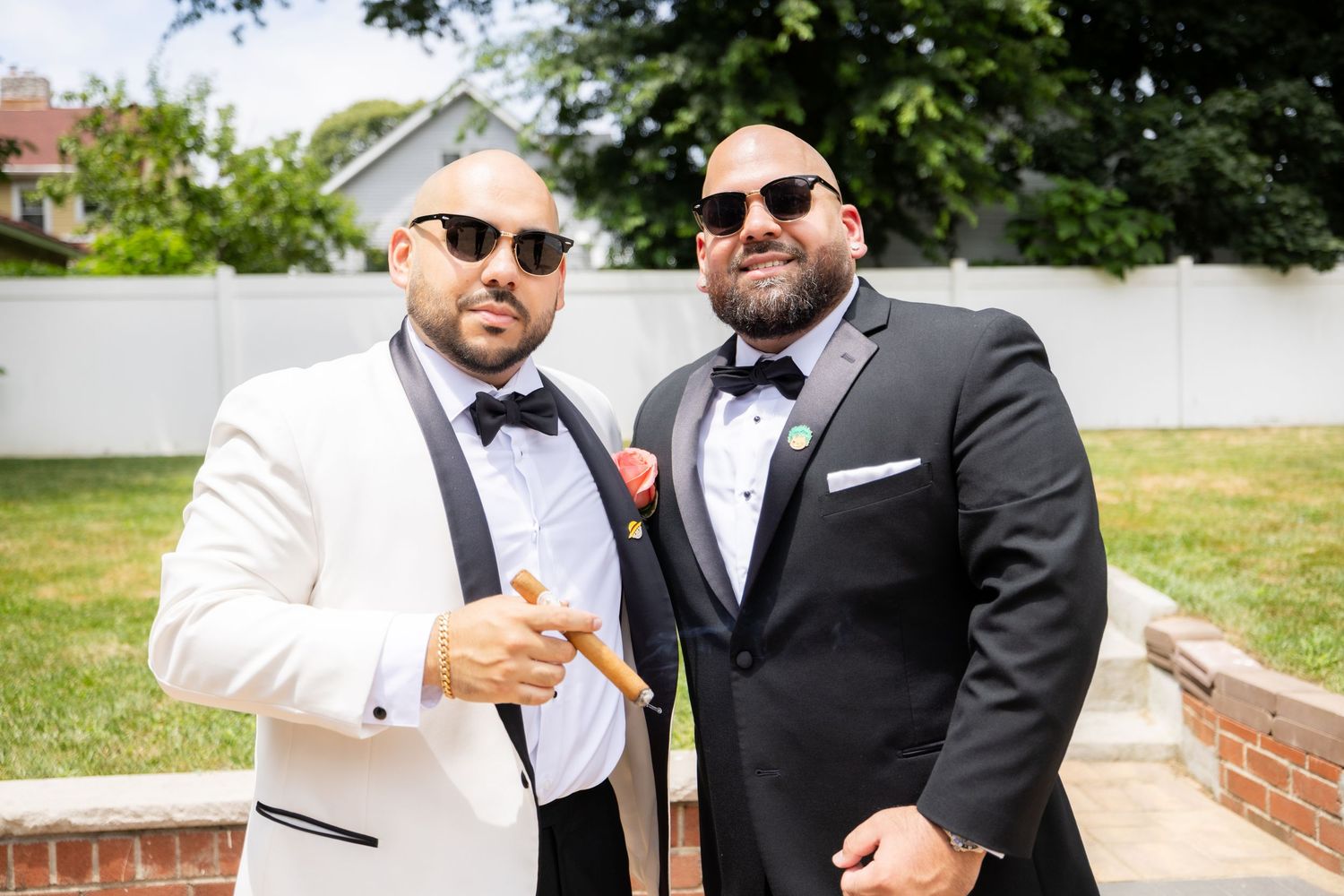 Two men in formal tuxedos, one in white and one in black, pose together outdoors wearing sunglasses.