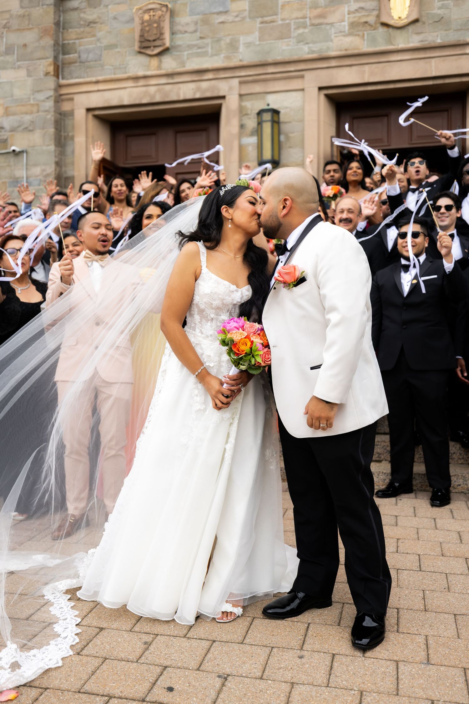 Wedding party celebrates outside a stone church with the bride in a flowing white gown.