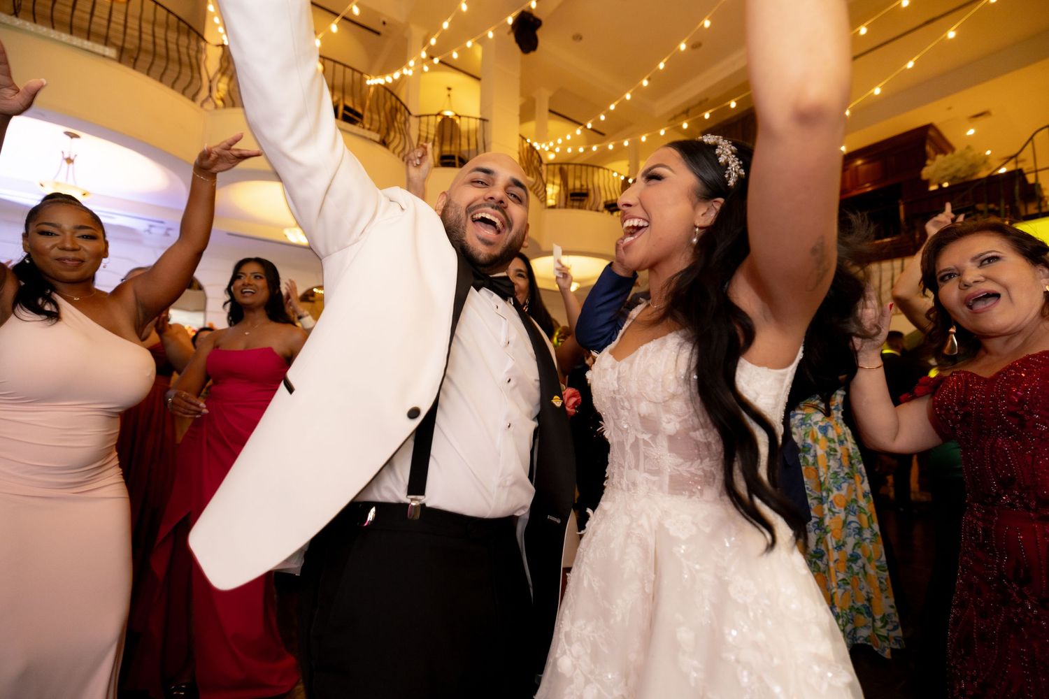Joyful wedding celebration with guests dancing under warm lighting in an ornate ballroom setting.