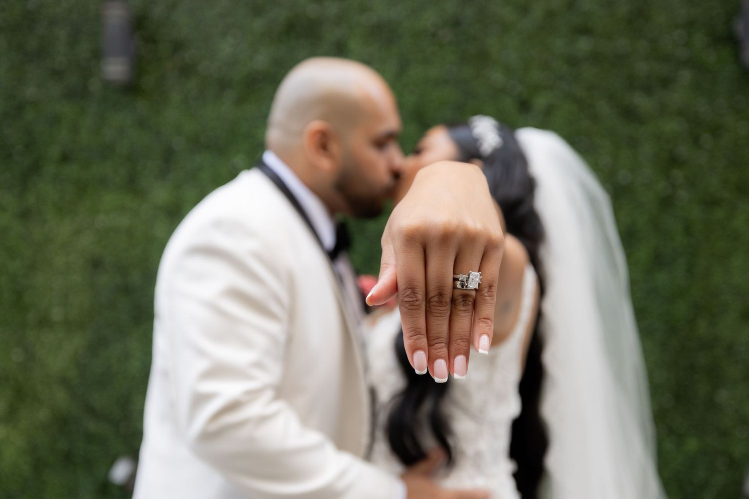 A close-up shot of a bride's hand showing her engagement ring while the couple shares a kiss in the background.