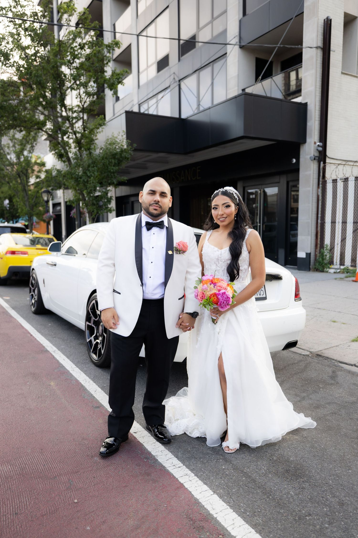 A couple poses by a white luxury car on their wedding day, with urban architecture in the background.