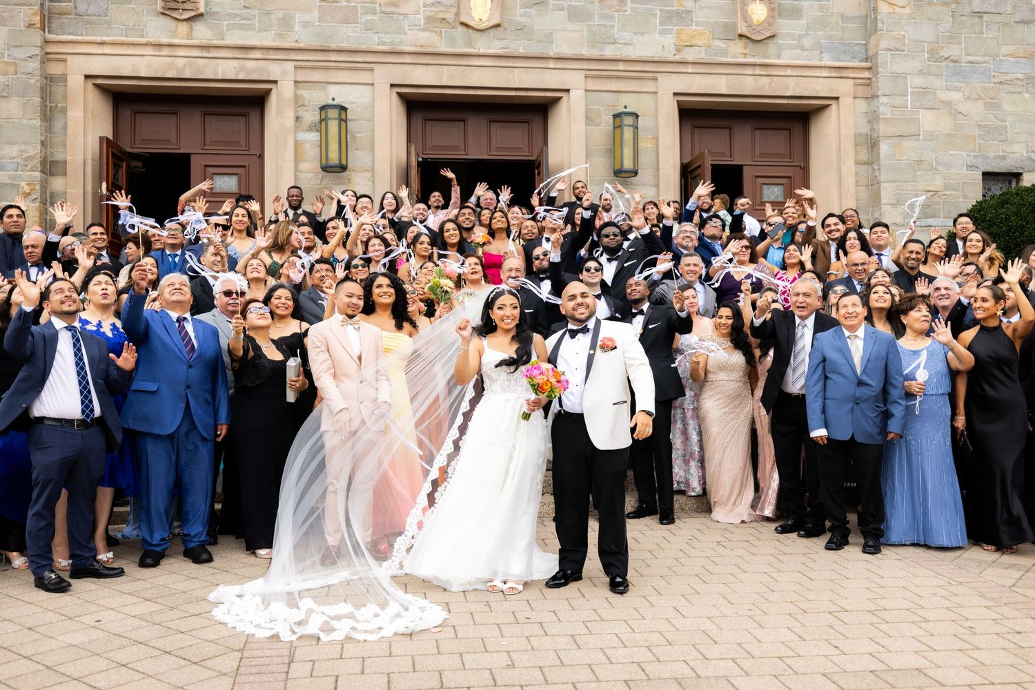Large wedding party and guests gathered outside stone church building for group photo celebration.
