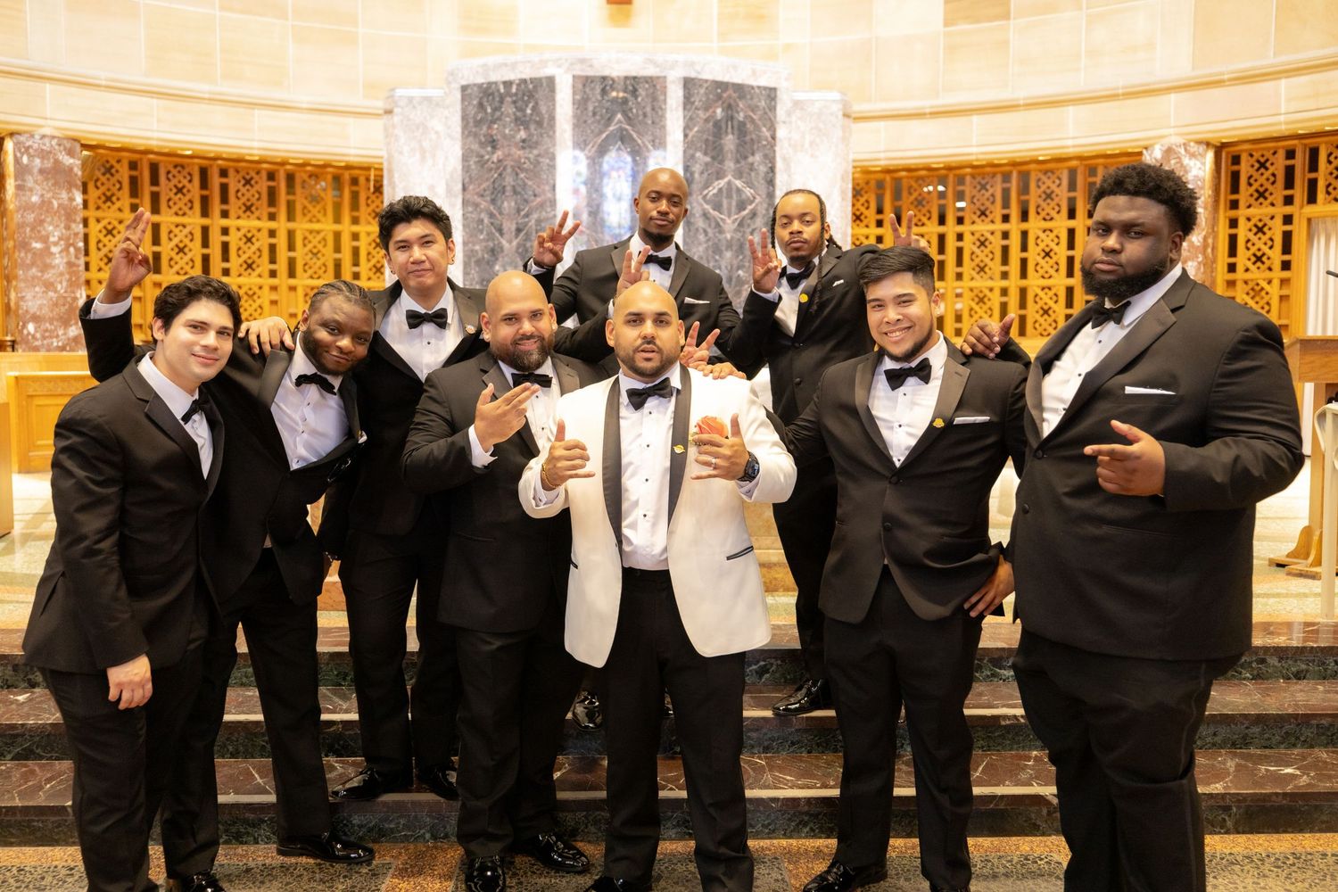 Wedding party in black tuxedos and formal attire poses together in an elegant venue with ornate gold decor.