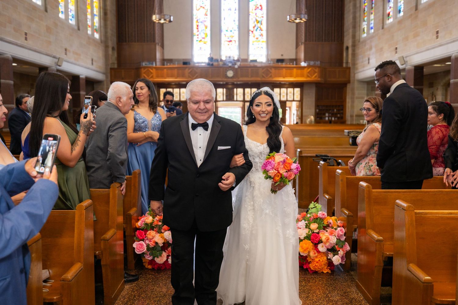 Wedding ceremony guests stand in wooden church pews adorned with colorful floral arrangements and stained glass windows.
