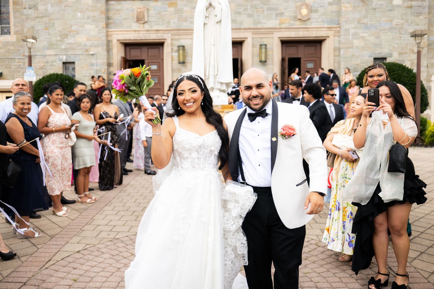 Wedding party celebrates joyfully outside a stone church building during a summer ceremony.