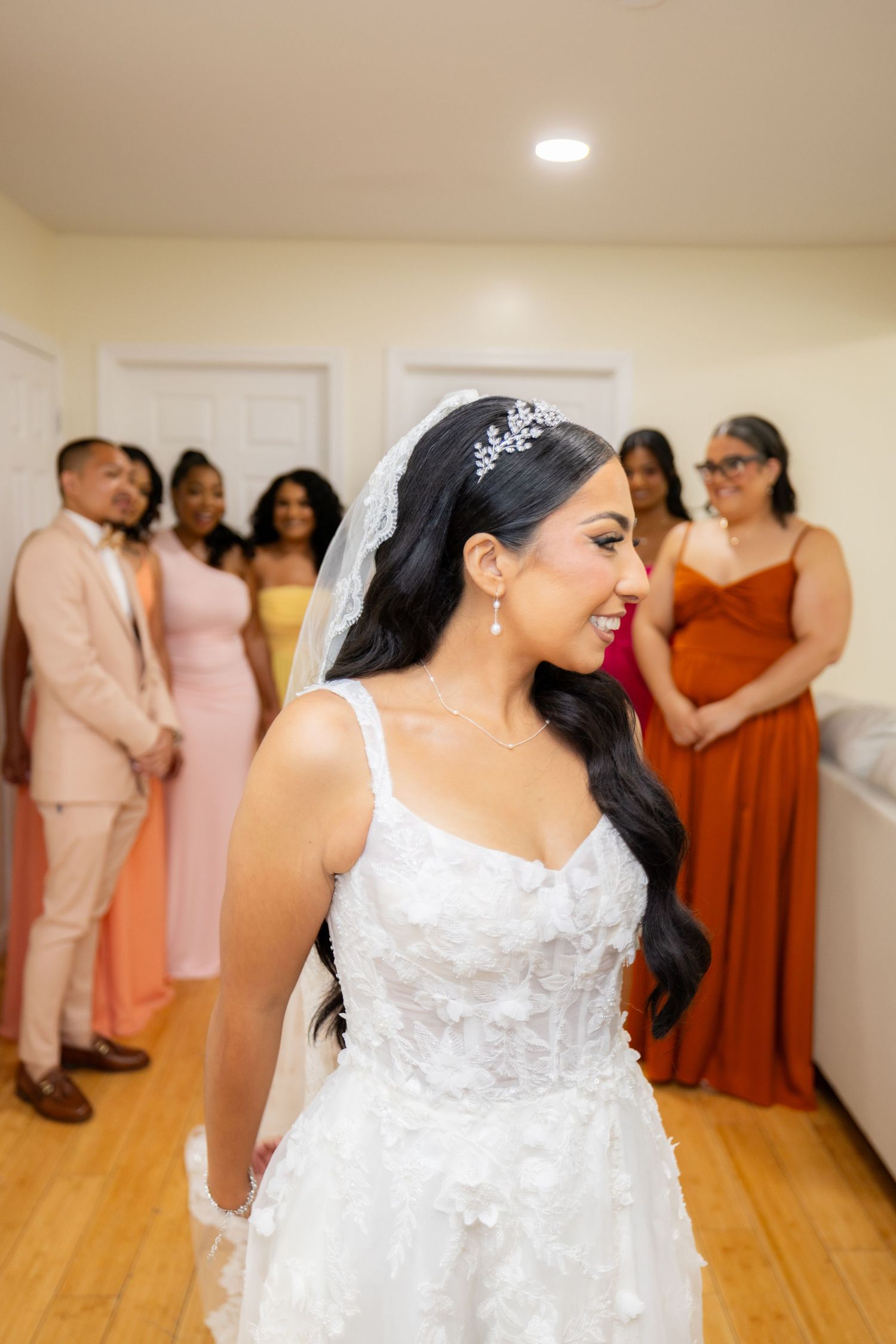 Wedding party members gather in a room with hardwood floors wearing rust orange and blush pink attire.