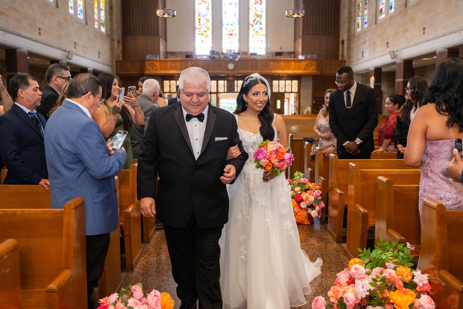 Wedding ceremony guests gather in a church with wooden pews decorated with bright floral arrangements and stained glass.