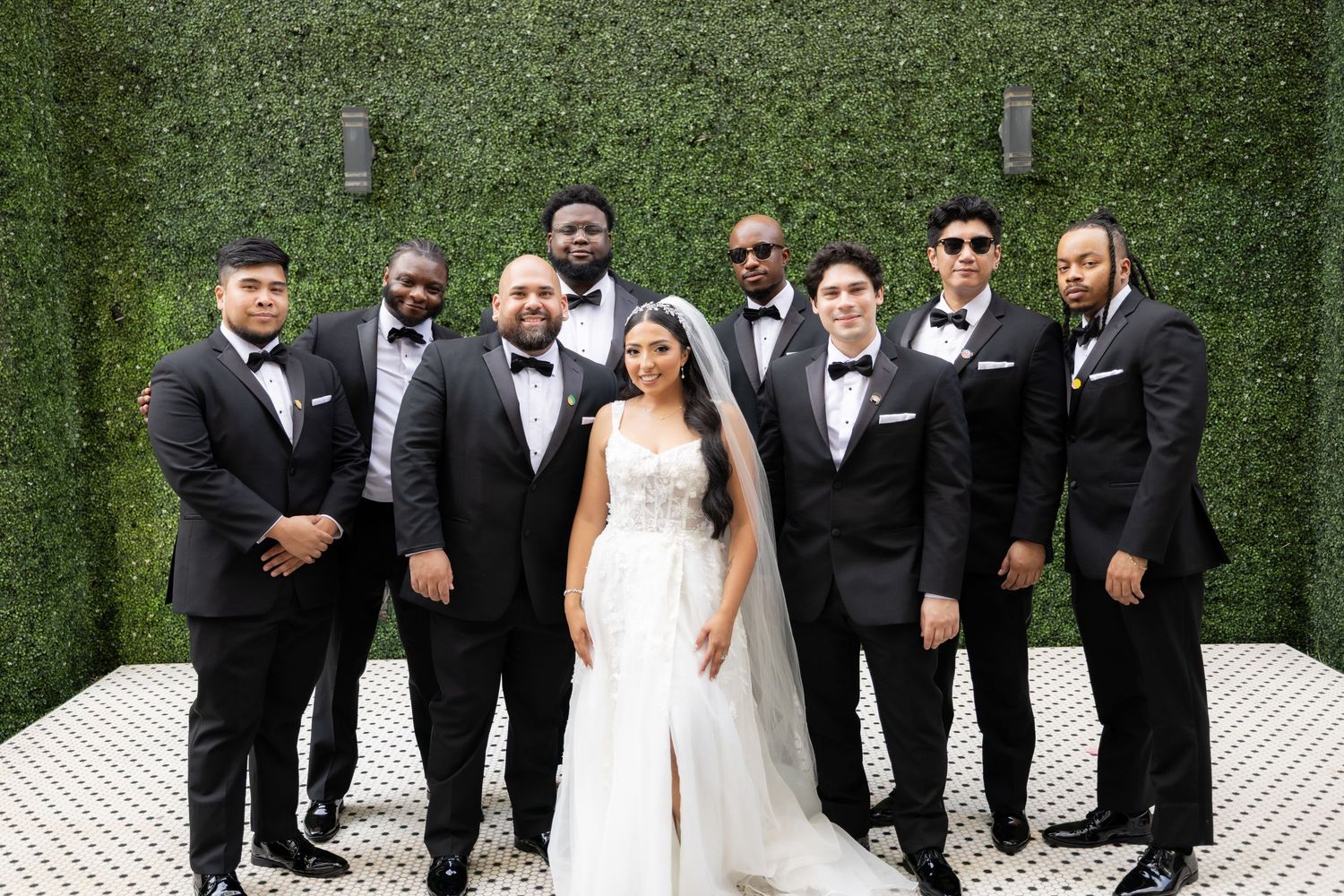 Wedding party poses outdoors against a lush green hedge wall in formal black and white attire.