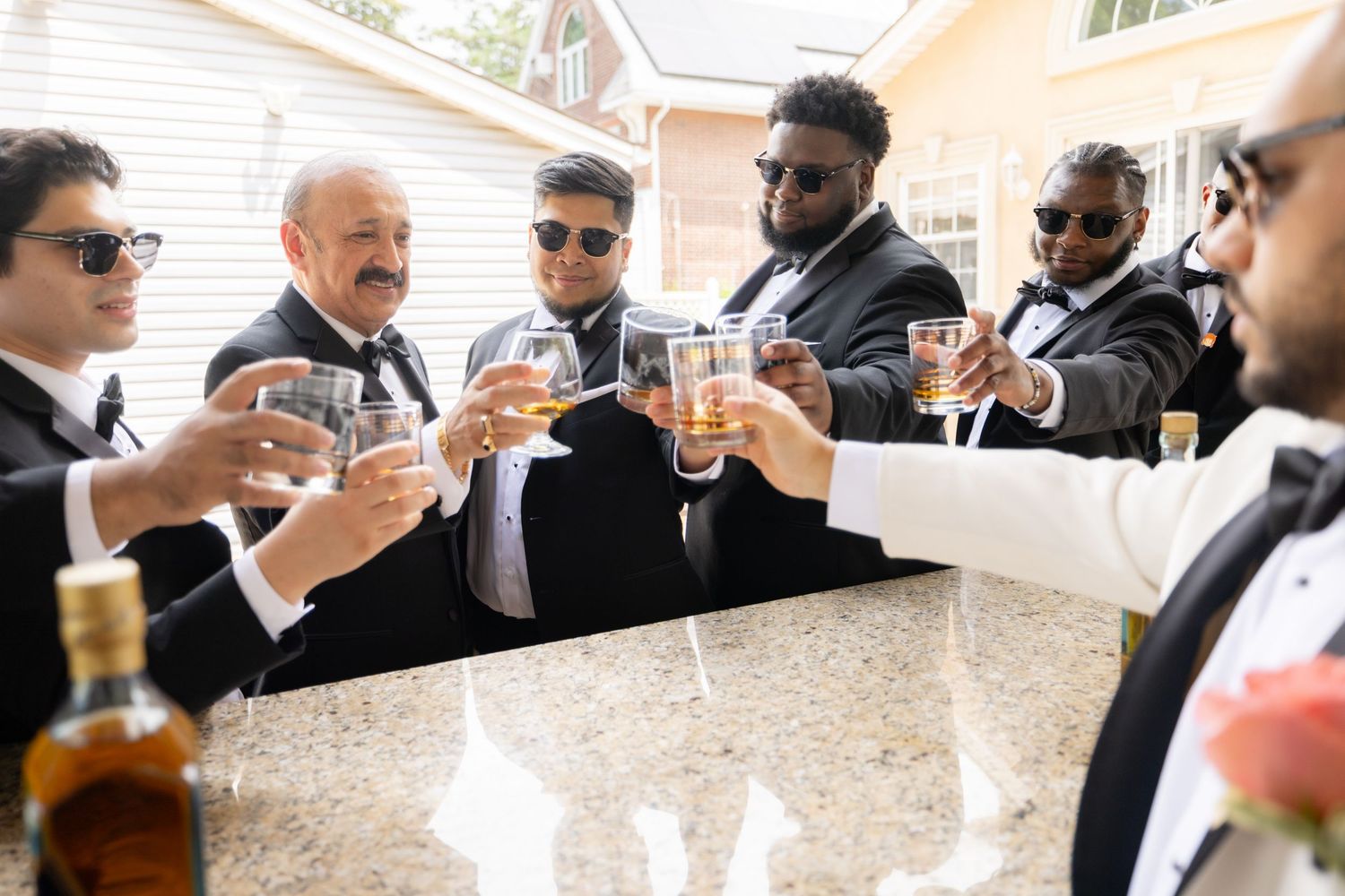 A group of groomsmen in black suits toast with glasses around a white marble countertop.